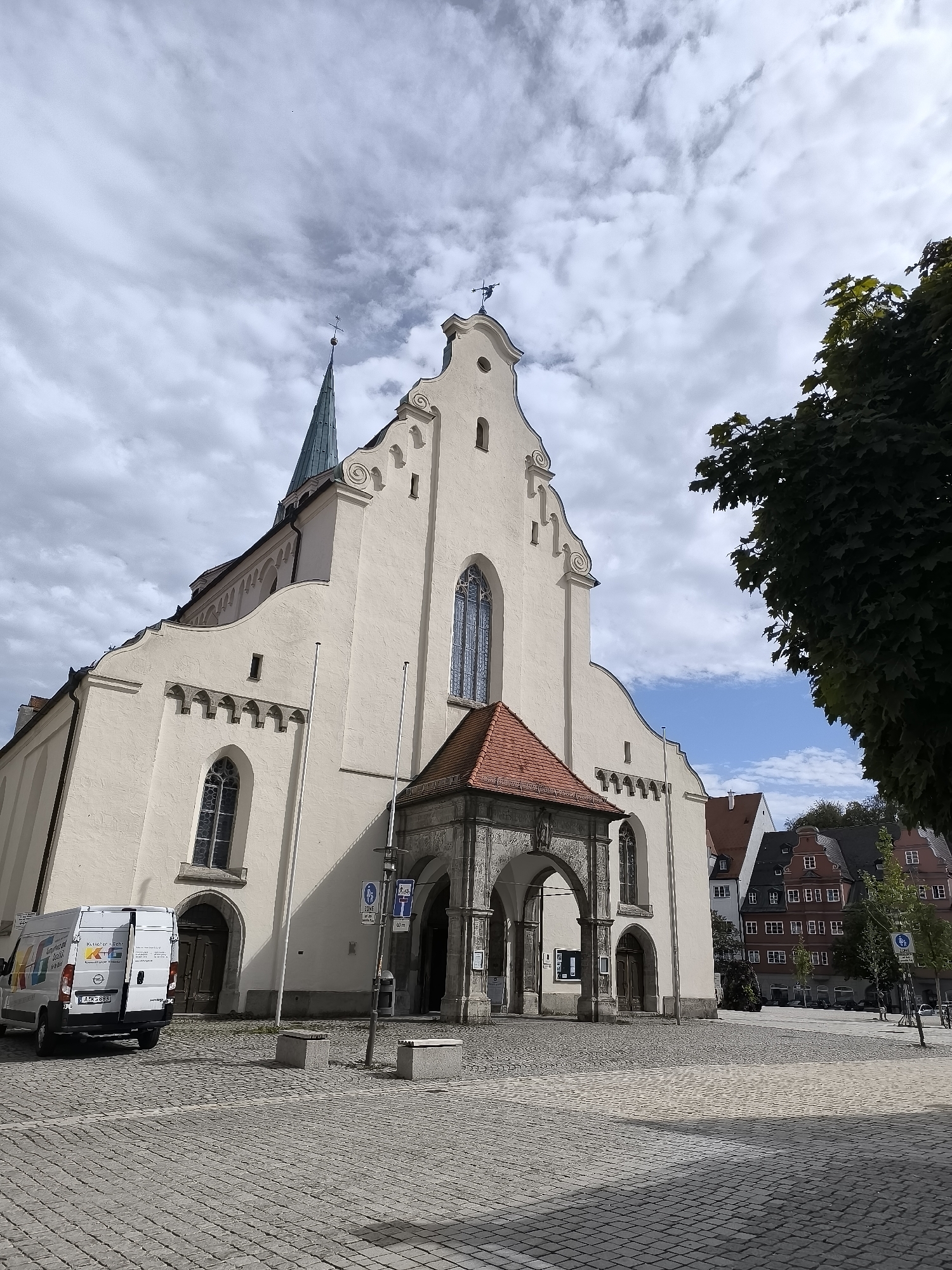 The image shows the exterior of St. Mang's Basilica in Kempten (Allgäu), Germany, captured from a slightly low-angle perspective. The church facade is predominantly cream-colored with a distinct green spire. The foreground is paved with cobblestones, and a white commercial van with the license plate "A-KS 883" is parked on the left. The van has colorful branding and "Opel" lettering. Several stone benches are scattered on the cobblestone plaza. The entrance to the basilica is partially covered by an arched red-tiled roof supported by stone pillars. In the background, traditional German buildings with red tiled roofs and white facades are visible, along with a large green tree on the right side of the frame. The sky is partly cloudy with patches of blue. No people are visible in the scene.