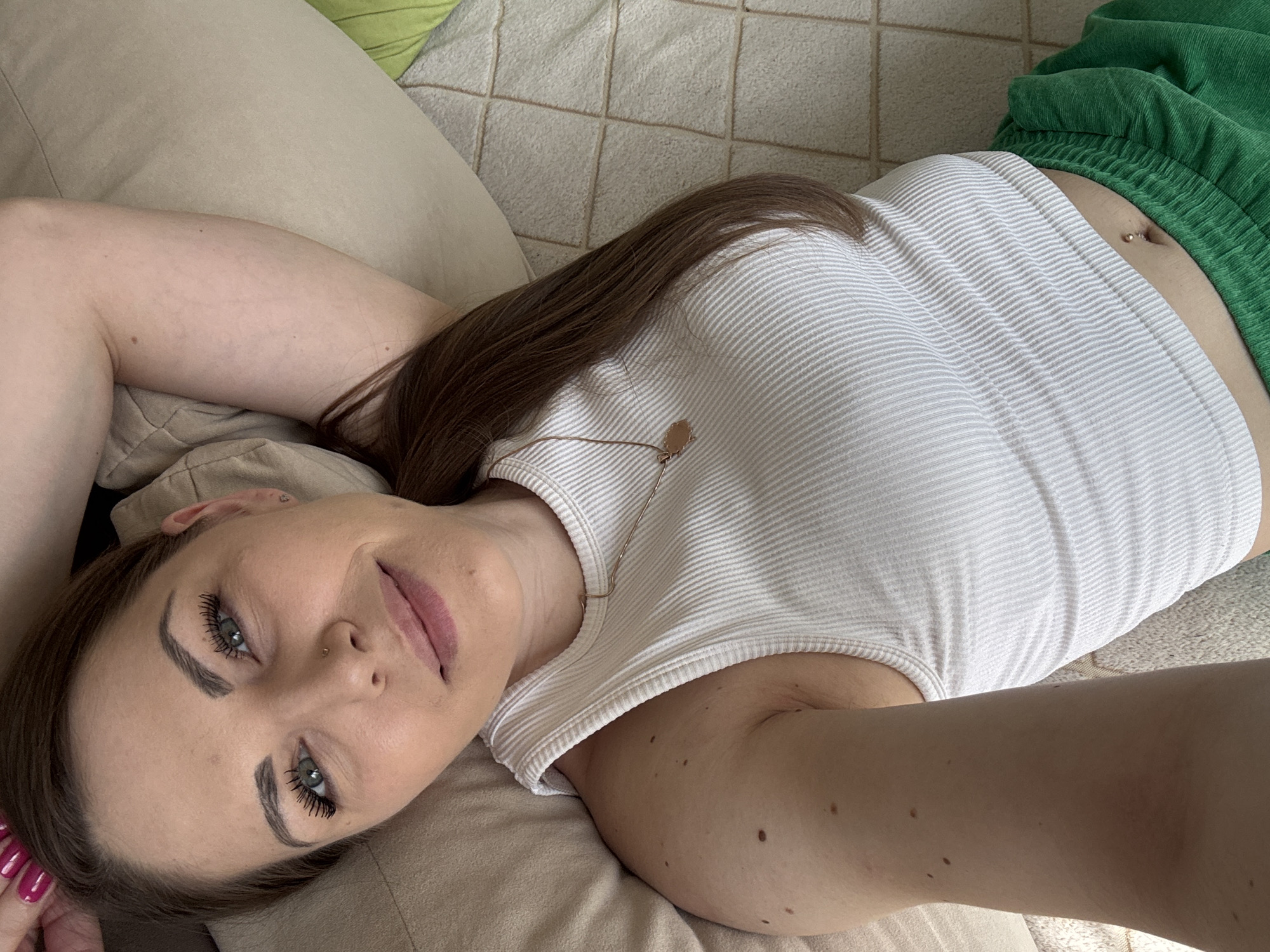 A woman lies on a beige-colored sofa with green cushions. She is wearing a white ribbed tank top and green sweatpants, and has a belly button piercing. She has brown hair and is wearing makeup. The background is a beige tiled floor. The photo was taken indoors. The location in the city of Раздолье, Russia is not discernible from the image.