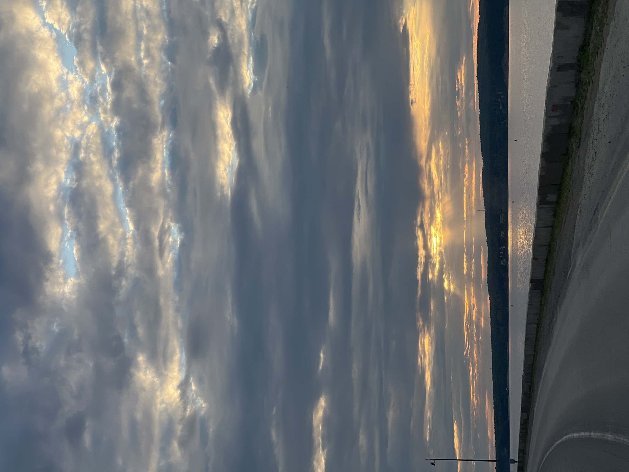 The image depicts a scenic view of the Kama River in Chaikovsky, Russia, during a sunset. The sky is filled with dramatic clouds, with rays of sunlight breaking through, casting a warm glow on the water. The foreground features a paved road curving along the riverbank, bordered by a concrete barrier. The opposite bank is lined with a dense forest. The overall atmosphere is serene and tranquil, showcasing the natural beauty of the region.