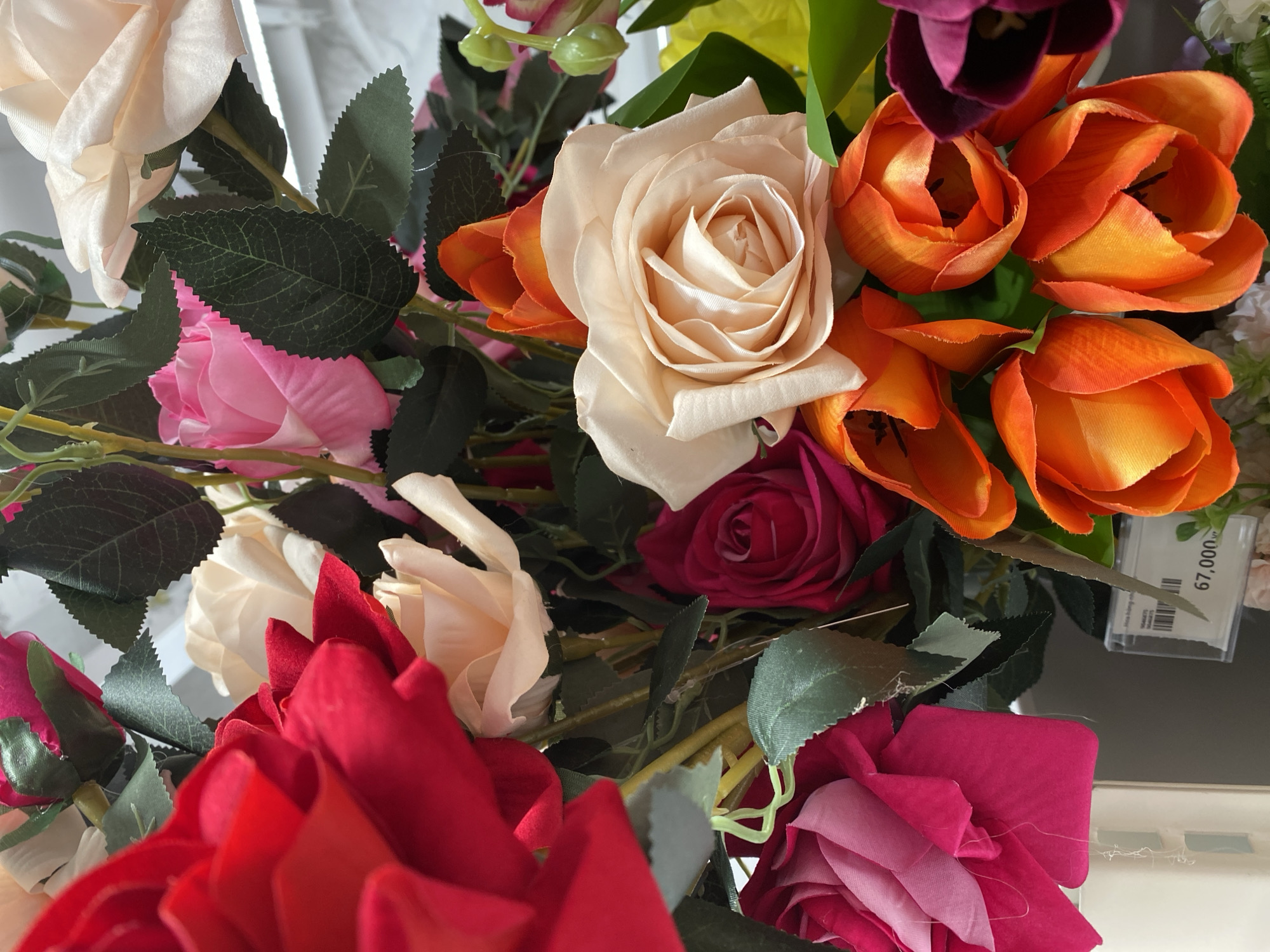 A close-up shot captures a vibrant arrangement of artificial flowers, predominantly roses and tulips, in various colors including red, pink, cream, and orange. The flowers are densely packed, creating a lush and textured bouquet. Some of the roses are fully bloomed, revealing intricate petal formations, while others are in bud form. The tulips, with their distinct cup-shaped blooms, add a splash of fiery orange. Dark green leaves are interspersed throughout the arrangement, providing a natural contrast to the colorful petals. In the lower right corner, a small price tag is visible, indicating a price of "67,000" and the currency "VND," suggesting the flowers are for sale in a retail setting in Vietnam. The background is softly blurred, hinting at an indoor environment, possibly a flower shop or a market stall in Đà Nẵng. There are no people visible in the frame.