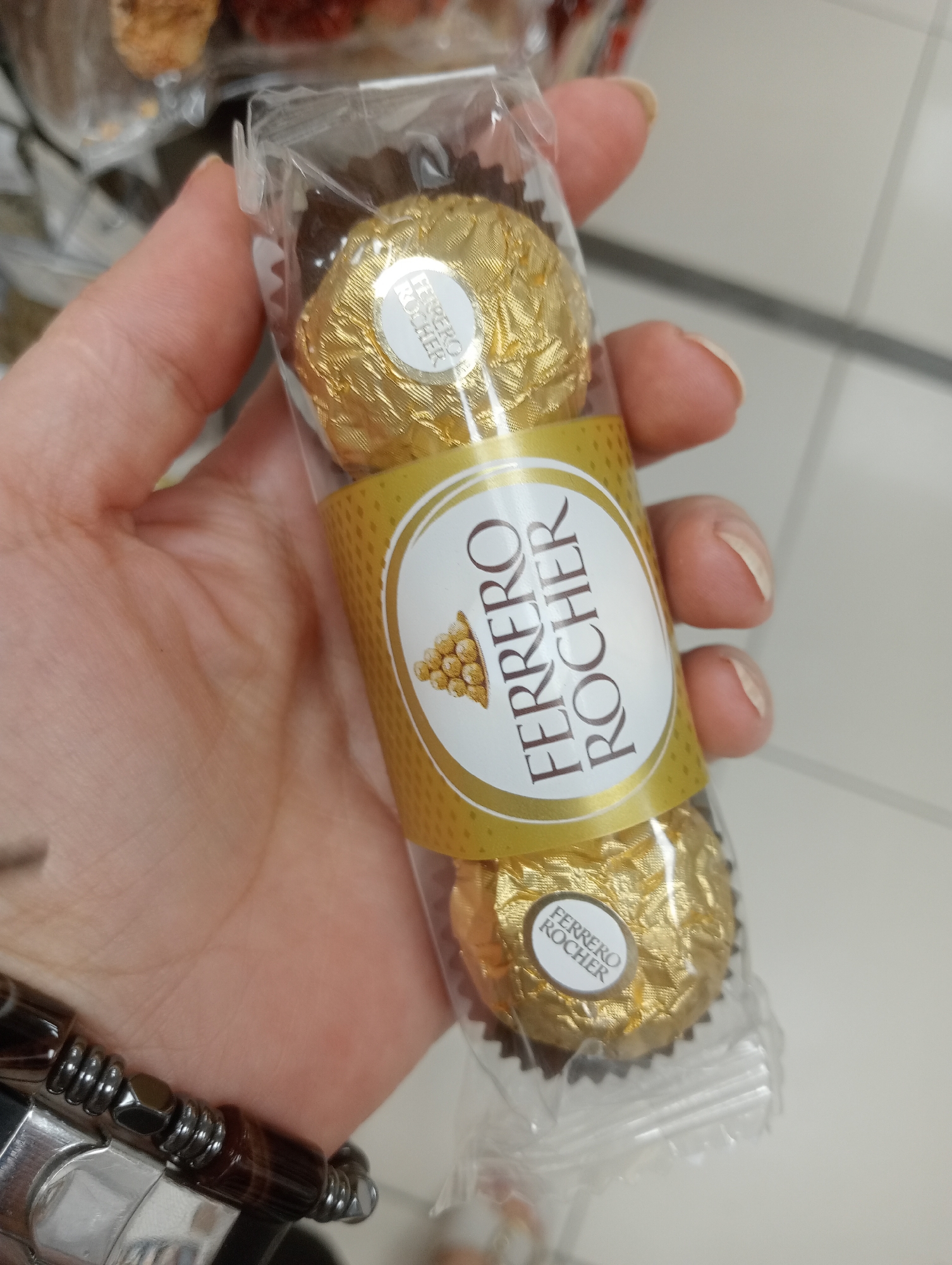 A hand holding a package of three Ferrero Rocher chocolates is shown. The chocolates are individually wrapped in gold foil. The package has a clear plastic wrapper and a gold band with the Ferrero Rocher logo. The background is blurred and appears to be an indoor setting, possibly a store. A watch or bracelet is visible on the hand. The image is taken from a close-up perspective.