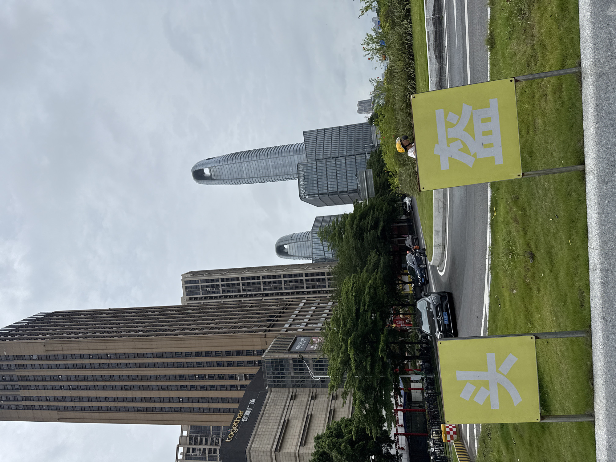 An overcast, daytime exterior shot in Haizhu District, China, captures a street scene dominated by modern high-rise architecture. In the foreground, two bright yellow signs with large white Chinese characters stand on metal poles, one displaying "头" and the other "篮". A person wearing a yellow helmet is visible in the background, seemingly riding a scooter. The signs are situated on a grassy area with a concrete border, adjacent to a curved road where several vehicles are present, including a black Mercedes-Benz sedan. Behind the road, lush green trees line the median, and further in the distance, several contemporary skyscrapers pierce the sky. One prominent building features a distinctive curved, open top, characteristic of modern architectural designs. Another tall, brown building with a grid-like facade is also visible to the left. Signs on a building to the left read "together" and "保时广埸". The overall scene depicts an urban environment with a mix of commercial buildings, traffic, and greenery.
