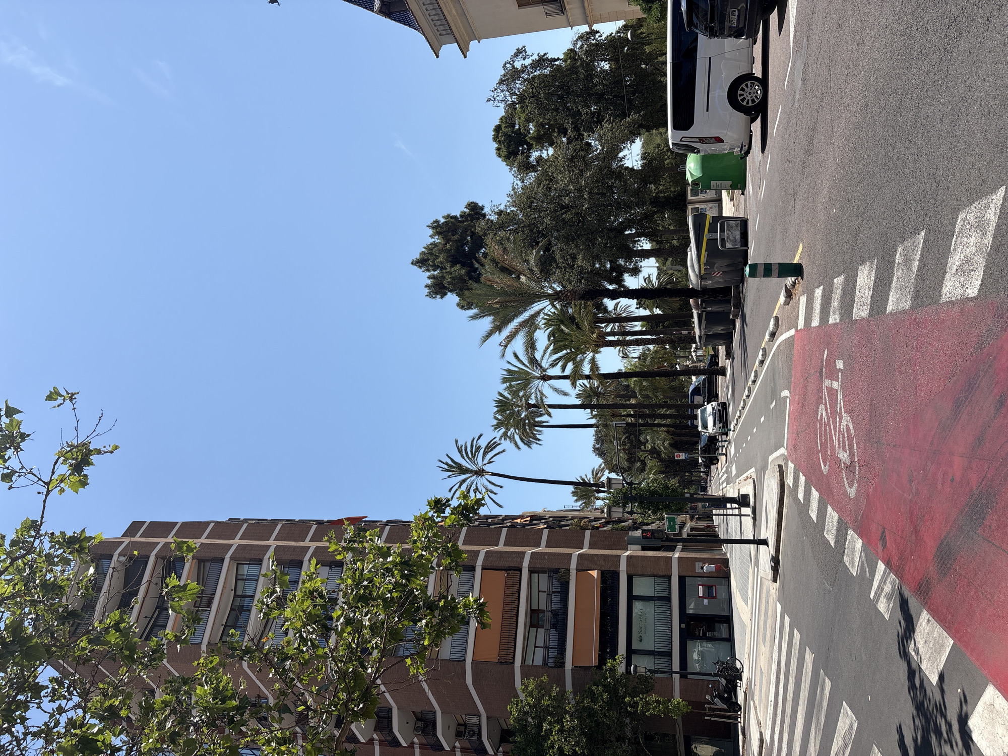 The image depicts a street scene in Valencia, Spain, on a clear, sunny day. The perspective is from street level, looking down a wide road lined with palm trees and buildings. In the foreground, a red-painted bike lane is visible, marked with a white bicycle symbol. A pedestrian crossing with white stripes is also prominent.
Several vehicles are parked or driving on the road, including a white van and a dark-colored car. Green and gray trash bins are positioned along the sidewalk. The buildings flanking the street are modern in style, with several stories and balconies. One building on the left has a distinctive facade with a patterned brick exterior and numerous windows. Trees with lush green foliage partially obscure the view of some buildings, suggesting a vibrant, urban environment with ample greenery.
The clear blue sky indicates it is daytime, likely late morning or early afternoon given the bright sunlight casting shadows. The overall atmosphere is calm and ordinary, capturing a typical urban streetscape. No people are clearly visible, contributing to a sense of quietness. There is a sign on the white van with text that is difficult to read. One of the shop windows shows the text "Santander".
