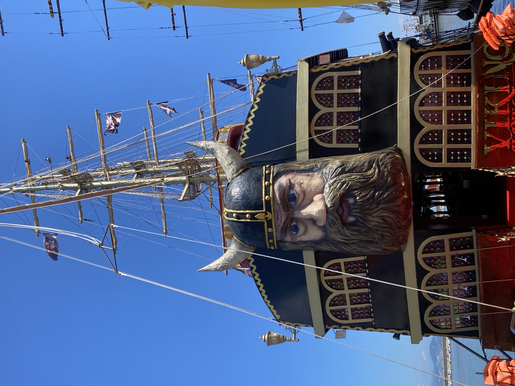 Captured from a low angle, the image showcases a Viking-themed ship in Alanya, Turkey. The ship's figurehead is a large, ornately carved head wearing a horned helmet. The ship's structure includes windows with latticework, a mast with rigging, and pirate flags. The sky is clear and blue. In the background, the sea meets a distant cityscape. Life vests are visible on the ship.