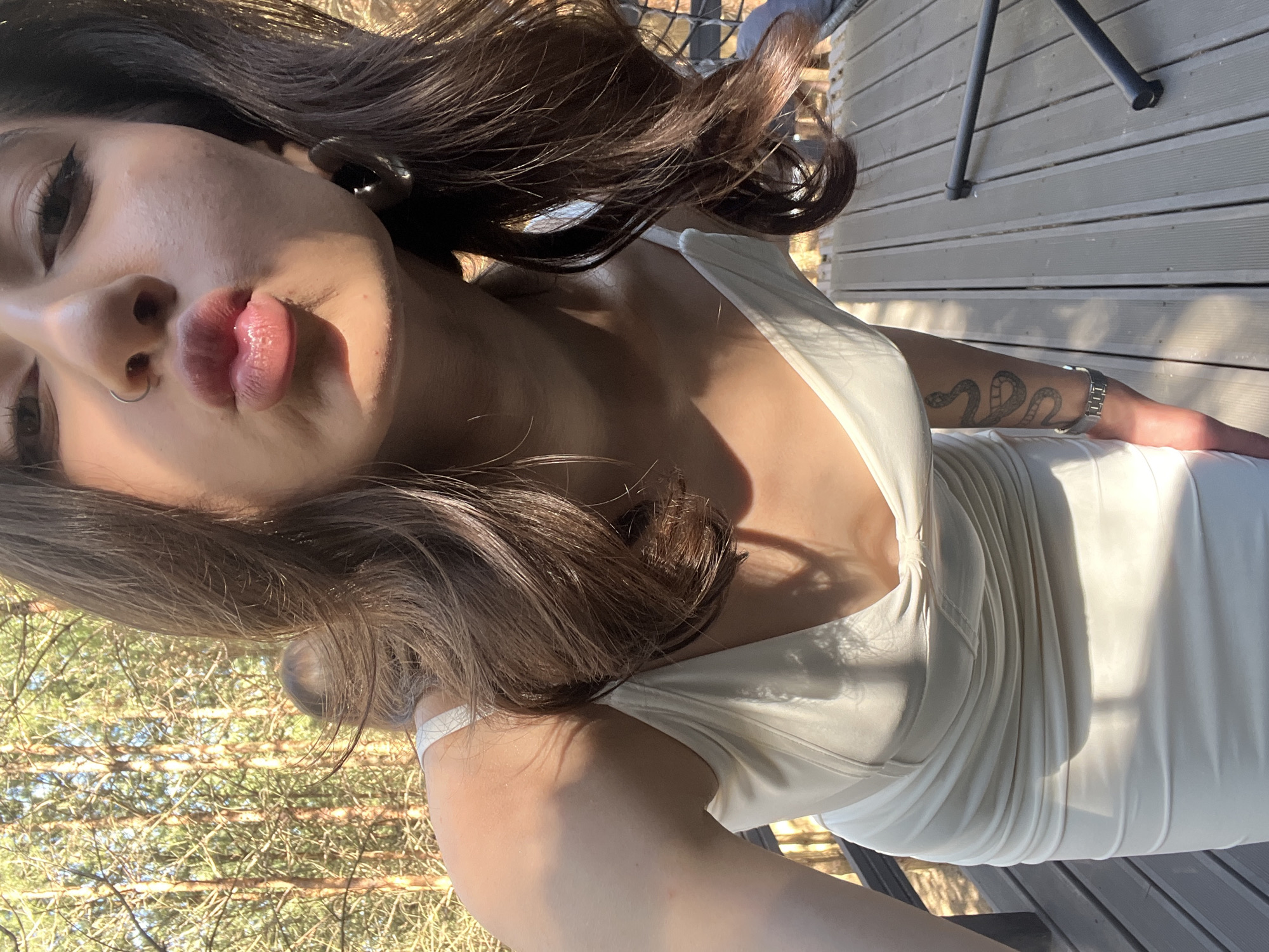 In Ivachiv Horishnii, Ukraine, a young woman is captured in a selfie on a wooden deck outdoors, surrounded by trees. She is wearing a cream-colored dress with thin straps and a draped neckline. Her lips are puckered, as if for a kiss. A silver hoop earring is visible in her right ear, and a nose ring is on her left nostril. Sunlight casts shadows across her skin and the dress. Her left arm displays a tattoo of a snake and a watch on her wrist. Another person is partially visible in the background, their back turned to the camera.