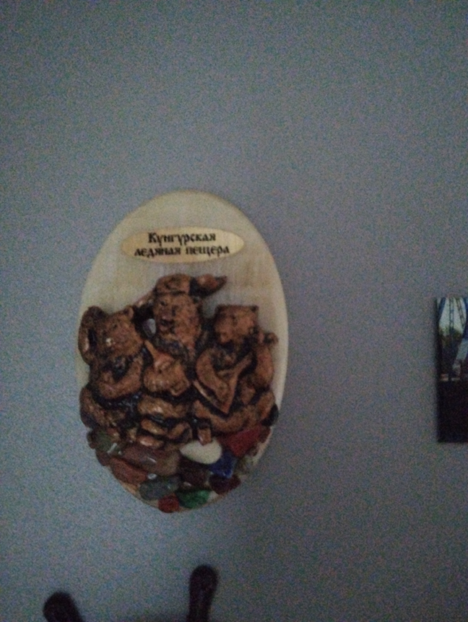 A close-up photograph depicts a mounted oval-shaped plaque with a sculpted depiction of three figures. A small rectangular placard above them is inscribed in Russian, "Kungurskaya ledyanaya peshchera", translating to "Kungur Ice Cave". The sculpted figures are brown and appear to be constructed from a textured material. The base of the oval contains an array of colorful stones or beads. The plaque is mounted on a wall, seemingly a light gray or blue-gray. To the right of the plaque, a smaller, partially obscured image or object is also mounted on the wall. Below the plaque, there is a blurred object that appears to be an object with knobs, likely a piece of furniture. Given the inscription referencing the Kungur Ice Cave, this photograph was likely taken in or around the city of Kungur, not Krasnogorskoye, Russia. There is no indication of Krasnogorskoye in this image.