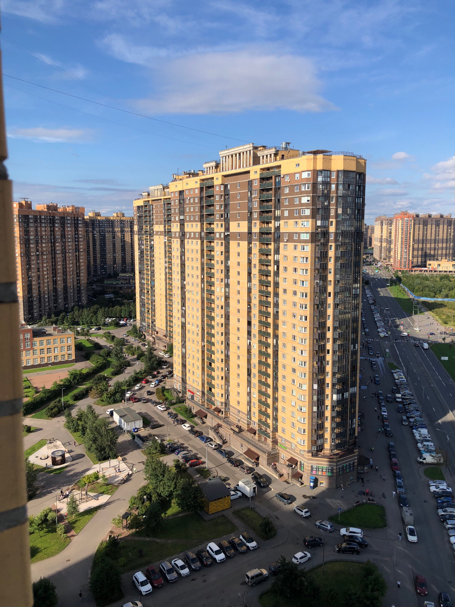 The image shows a residential area in Lavriki, Russia, on a sunny day with scattered clouds. The scene is dominated by a large, modern apartment building with a beige and dark brown facade, featuring numerous balconies and windows. In the foreground, a portion of another building, possibly made of brick, is visible.
Several other tall apartment buildings are visible in the background, some with orange and beige facades, suggesting a high-density urban development. A road runs alongside the main apartment building, with many cars parked on the side and a few moving. A sidewalk is present along the road, with some pedestrians walking.
The area surrounding the buildings includes green spaces with trees, lawns, and paved pathways. There are also playground areas with children's equipment and seating. The overall impression is of a well-maintained, contemporary residential complex. The sky is a clear blue with streaks of white clouds.