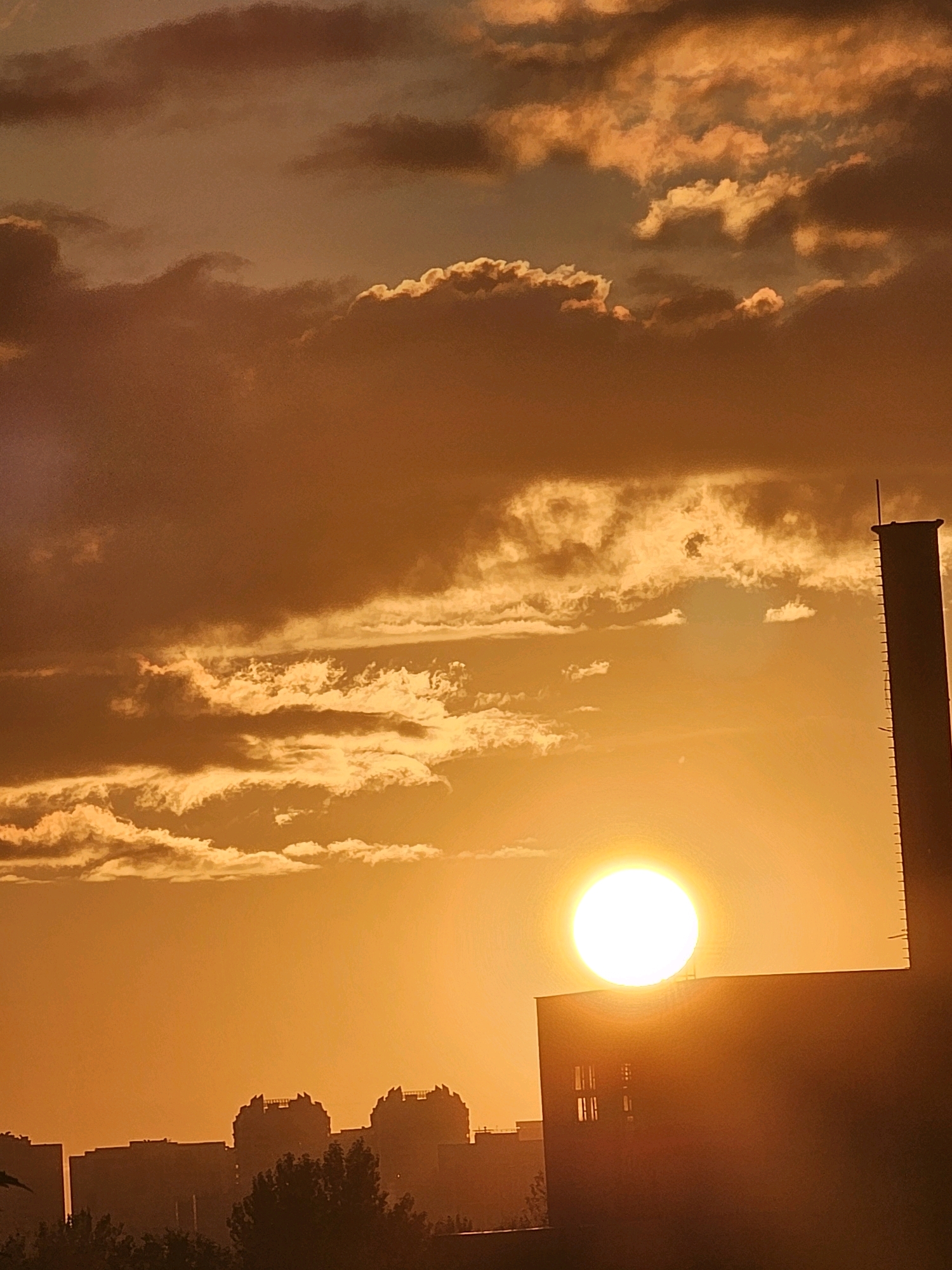 The image depicts a sunset over the city of Khimki, Russia. A large, bright sun is positioned near the horizon, casting a warm, orange glow across the sky and illuminating scattered clouds. In the foreground, a dark, silhouetted building occupies the lower right portion of the frame, with a tall, slender structure, possibly a chimney or tower, rising from its roof. On the left, a silhouetted skyline of buildings and trees suggests a residential or urban area. The overall scene is characterized by the dramatic lighting of the setting sun, creating a serene yet imposing urban landscape.