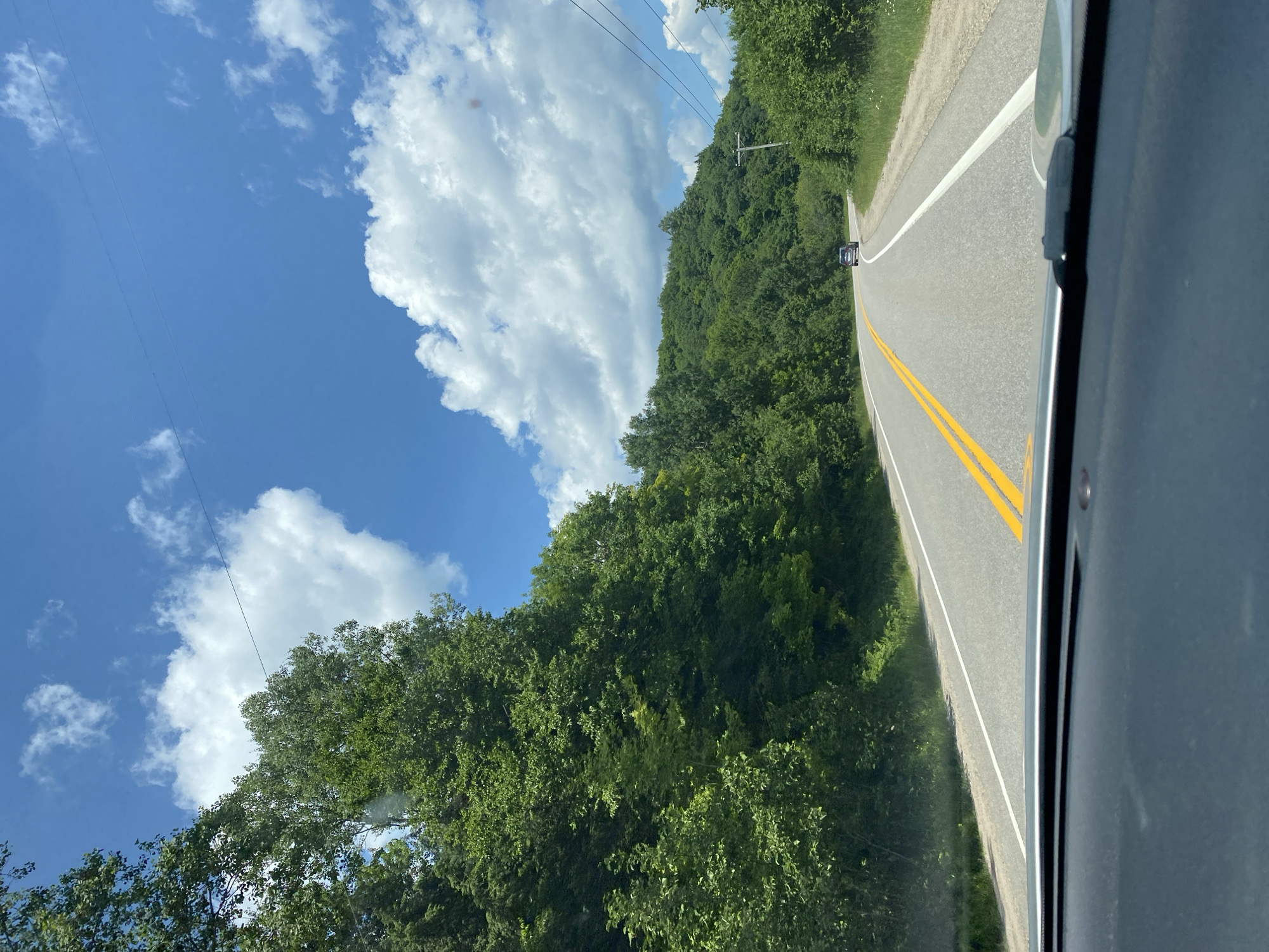 The image depicts a point-of-view shot from a vehicle traveling on a paved road that curves to the right, surrounded by lush green trees and rolling hills under a partly cloudy blue sky. The road has a white line marking the right edge and double yellow lines in the center. A dark-colored SUV is visible in the distance, traveling in the same direction. Power lines run diagonally across the sky. The foreground shows the dashboard of the vehicle from which the photo is taken. There is no indication of the specific location within the city of Lac-Simon, Canada.