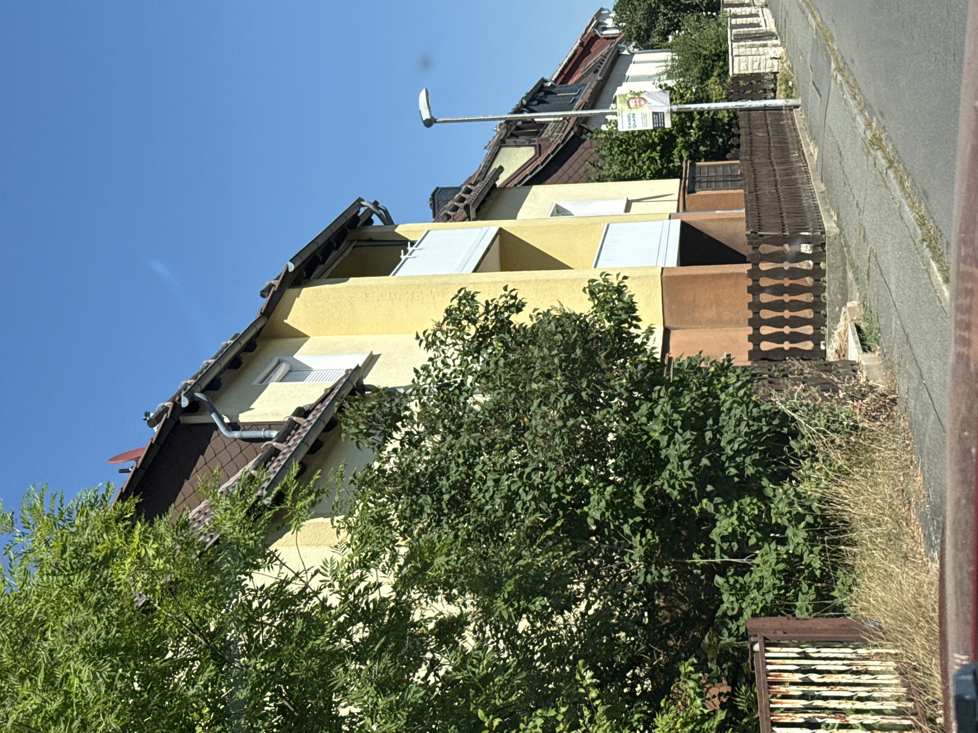 The image captures a residential street scene in Bad Sachsa, Germany, under a clear blue sky. The foreground shows the edge of a vehicle's dashboard and windshield, indicating the photo was taken from inside a car. A paved street runs along the right side of the image, bordered by a sidewalk.
Dominating the scene are buildings with yellow facades and brown tiled roofs, characteristic of German residential architecture. A prominent feature is a wooden fence with decorative spindles lining the front of one of the properties. Beyond the fence, overgrown dry grass and lush green trees and bushes add to the natural setting.
A street lamp stands tall near the sidewalk, and attached to it is a political poster featuring a smiling man named Daniel Quade. The poster has a light-colored background with text in a darker color. The buildings appear to be multi-story dwellings, with balconies visible on one of the structures. The overall impression is a quiet, sunlit day in a German town.