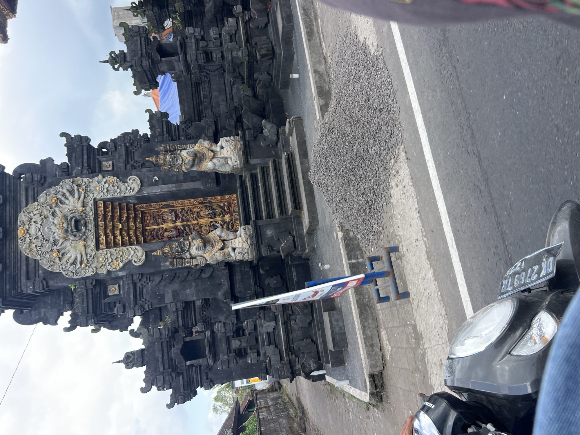 The image captures a street view in Abiansemal, Indonesia, featuring a traditional Balinese temple gate constructed of dark stone with intricate carvings. The gate has a large, ornate golden door, flanked by two warrior statues. In front of the temple, a pile of gravel is visible on the sidewalk, with a blue A-frame sign standing nearby. A gray motorcycle is in the foreground, with its license plate, "DK 2769 TZ," clearly visible. The street is paved, and a white line marks the edge of the lane. The sky is overcast with clouds. The scene appears to be a typical street in a residential or commercial area in Abiansemal.