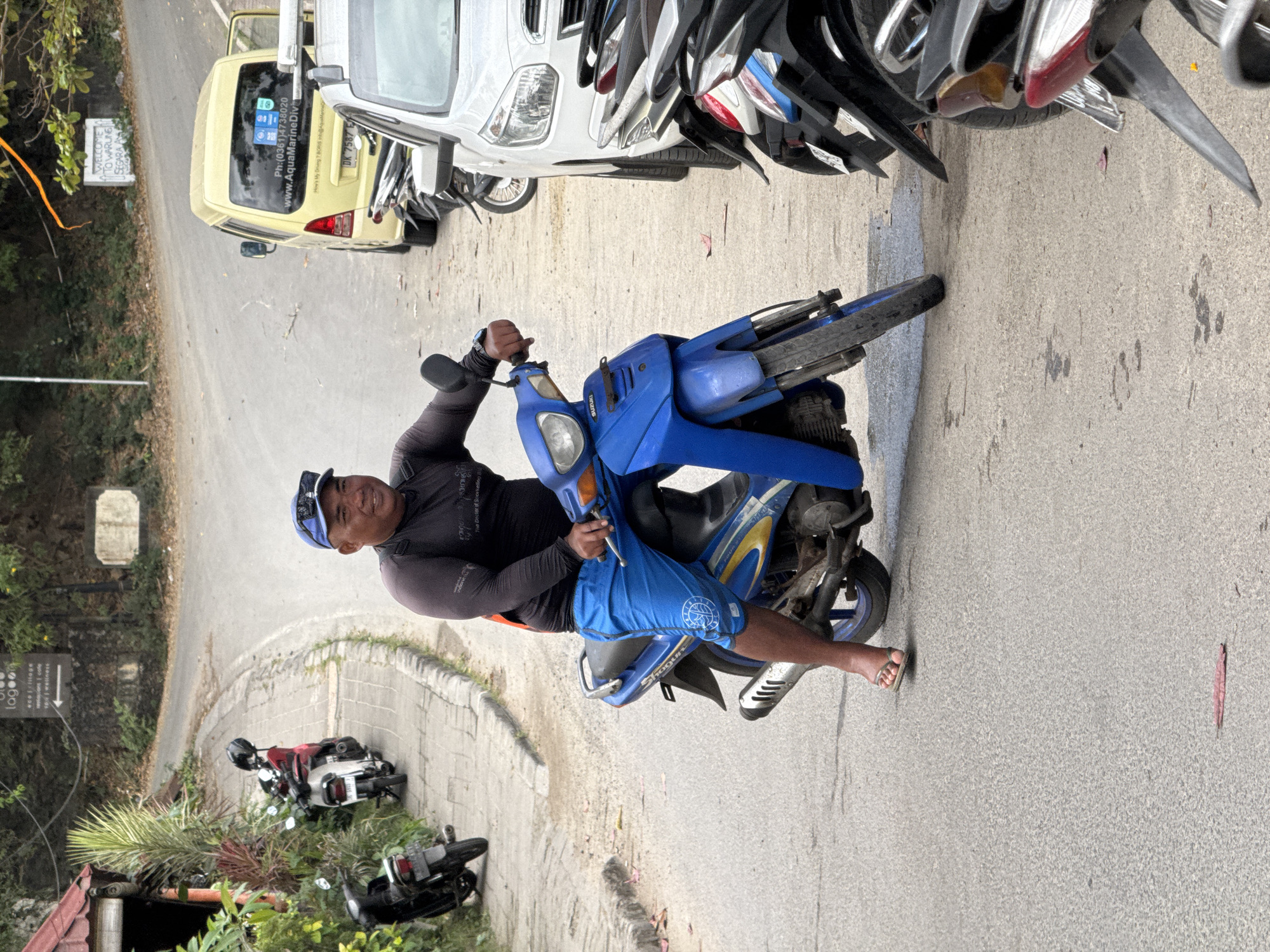 A man is sitting on a blue Suzuki motorcycle in Manggis, Indonesia. He is wearing a black long-sleeve shirt, blue shorts, and a blue baseball cap. He is looking at the camera and smiling. The motorcycle is parked on a paved road. In the background, there are other motorcycles, a white car, and a yellow van. There are also some trees and a sign in the background.