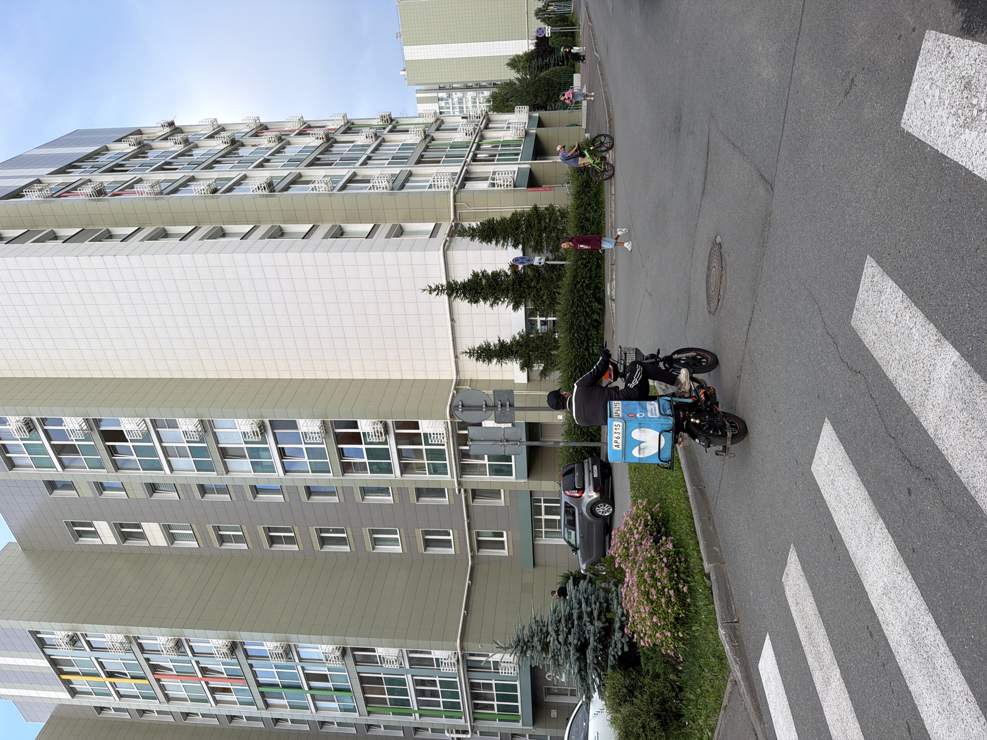 In a residential area, likely Kazan, Russia, a delivery person rides a scooter past a large, multi-story apartment building and a pedestrian crossing. The building is modern, with a light grey facade and many windows. The scooter has a blue insulated delivery box attached to the back, with the text "AP 6315" visible on it. The pedestrian crossing is marked with white stripes on the asphalt road. Several people are visible in the background, some on bicycles. The sky is clear and the lighting suggests it is daytime.