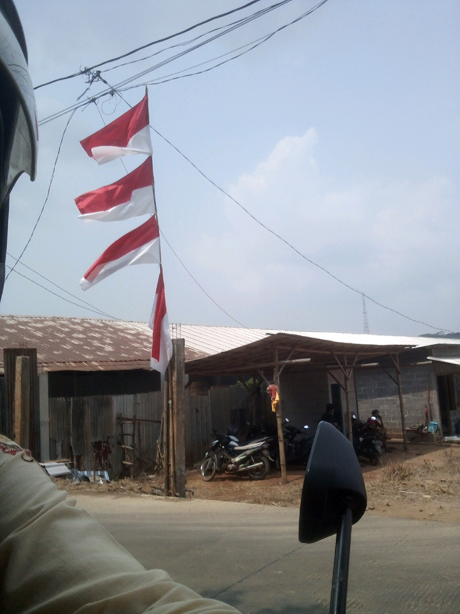 The image depicts a scene in Banyuputih, Indonesia, featuring several Indonesian flags (Sang Saka Merah Putih) flying from a pole against a pale blue sky with some wispy clouds. The flags are positioned in a vertical line, with the top flag appearing slightly larger and more spread out than the two below it, which are partially furled. In the foreground, a black rearview mirror of a motorcycle is prominently visible, along with a portion of the rider's beige clothing and helmet.
Behind the flags, a building with a corrugated metal roof supported by wooden beams is visible, suggesting a rural or semi-urban environment. To the right of this structure, another building constructed with gray cinder blocks is present, with a covered outdoor area in front of it. Several motorcycles are parked under this covered area, and two individuals can be seen, one sitting and the other standing. The ground appears to be a dirt road or pathway. Overhead, electrical wires crisscross the sky. The overall impression is one of a typical roadside scene in Indonesia, possibly adorned with flags for a national event or celebration.