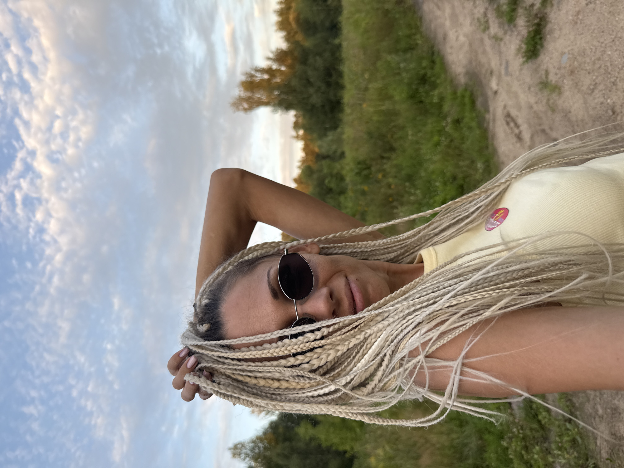 A woman with blonde braids and sunglasses is posing for a selfie outdoors. She is wearing a yellow top with a pink badge. The background shows a grassy field and trees under a cloudy sky. The image was taken in Yaroslavl, Russia.
