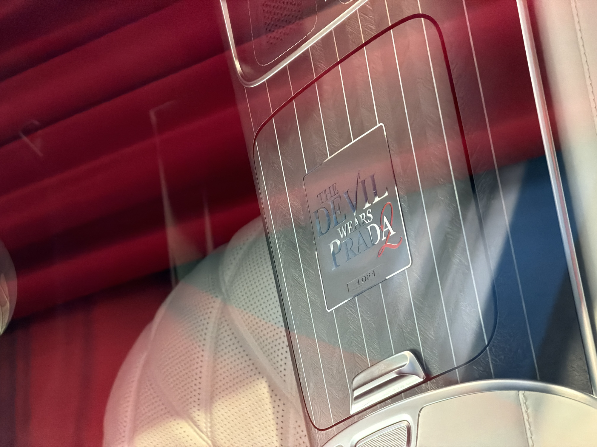The image shows a close-up of a car's interior, focusing on a metallic panel with horizontal lines. A rectangular emblem is affixed to this panel. The emblem displays the text "THE DEVIL WEARS PRADA" in stylized lettering, with "PRADA" in a bolder font and highlighted with a red accent. Below the main text, the number "1907" is visible, also in a stylized font. The car's interior features white perforated seating in the background and red fabric curtains or lining. The lighting suggests this scene is captured indoors, possibly in a showroom or a private collection, in Munich, Germany. There are no people visible in the frame.