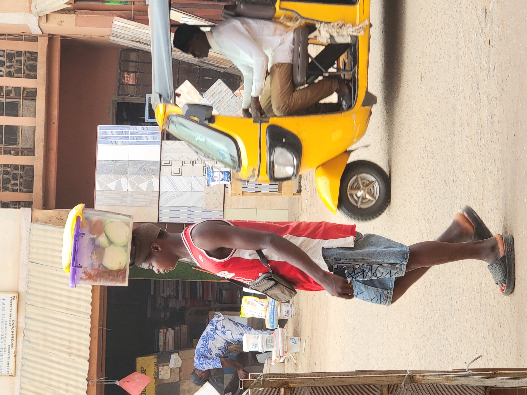 This vibrant image captures a bustling street scene in Maiburiji, Nigeria, under bright daylight. The primary subject is a young man walking from left to right, carrying a clear plastic container filled with sliced green fruit, likely guava, and a reddish spice mixture on his head. He is dressed in a red and white sleeveless jersey, denim shorts, and black slides, with a small black bag slung across his chest.
In the background, a bright yellow motorized tricycle, commonly known as a Keke Napep, is visible. A man in a white shirt and brown trousers is seated in the driver's seat. To the left, behind the walking man, various types of wall and floor tiles are displayed prominently, indicating a tile shop or construction material supplier. Another individual, partially obscured and wearing a patterned blue and white garment, is seen bending over near some products in front of the shops.
Visible objects include buckets labeled "EVA" and "EVA BOND," likely adhesive or other construction materials. A sign on a corrugated metal roof provides details for a business: "SIKENALISTE ELCARELCTICE OF A TYPES & GENELAL SUPPLIES AR 3 KANO RR, KAPUNA AD2 KANO RD, KADUNA 08036667098". Other text includes "JP", "thestos-freenoor Tiles", and "737" on a red item on a wall. The scene depicts a common daily activity of street vending alongside local commerce, reflecting the dynamic atmosphere of a Nigerian market street.