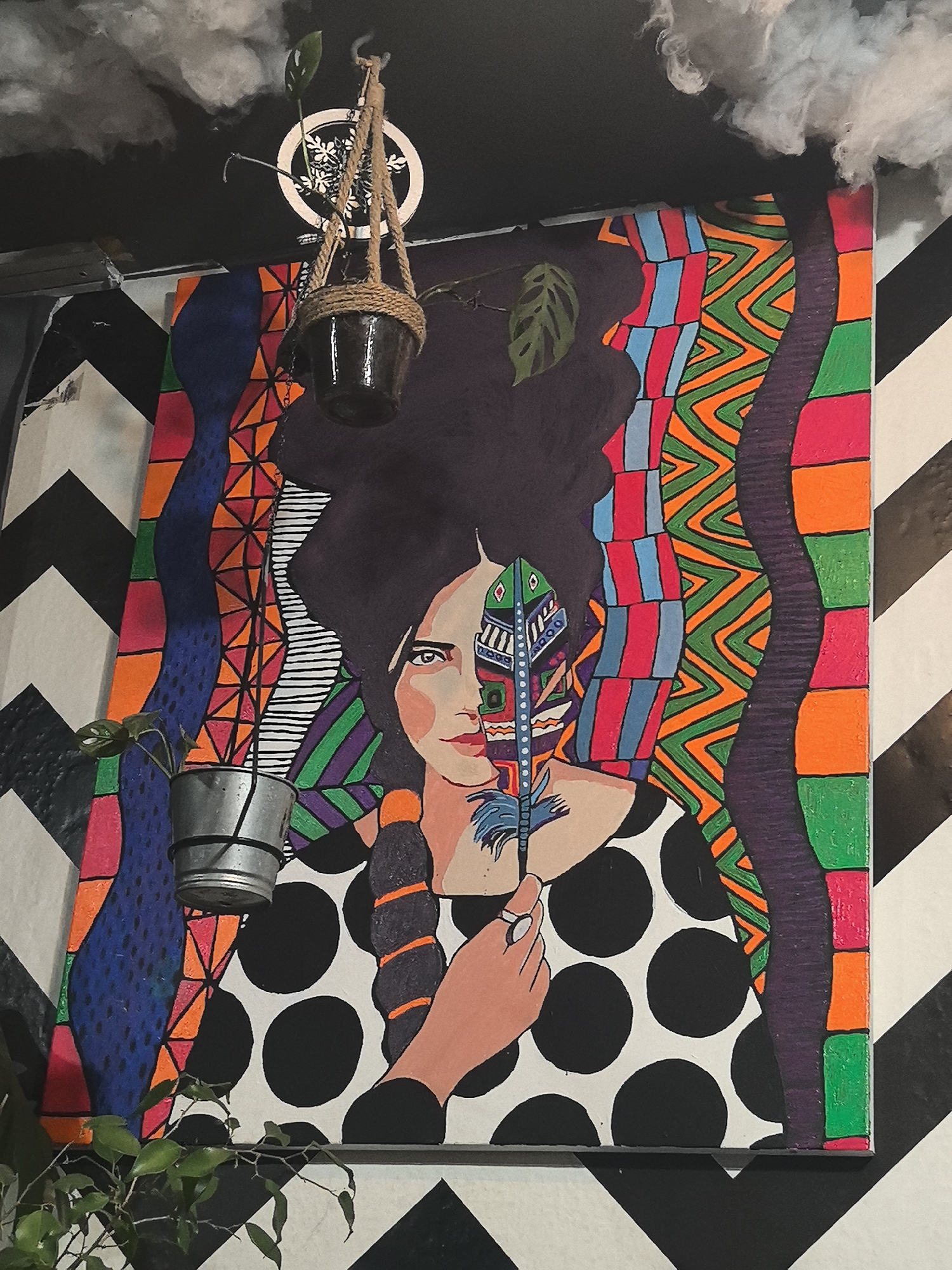 A framed, colorful painting is displayed on a wall with a black and white chevron pattern. The painting depicts a woman with dark hair, part of her face obscured by a feather. Her dress is white with large black polka dots. The edges of the painting are decorated with colorful, geometric patterns. A hanging planter with a small plant is positioned on either side of the painting. White cotton-like material is attached to the ceiling above the painting. The location in the city of Noviy, Russia, cannot be verified as this information is not discernible from the image.