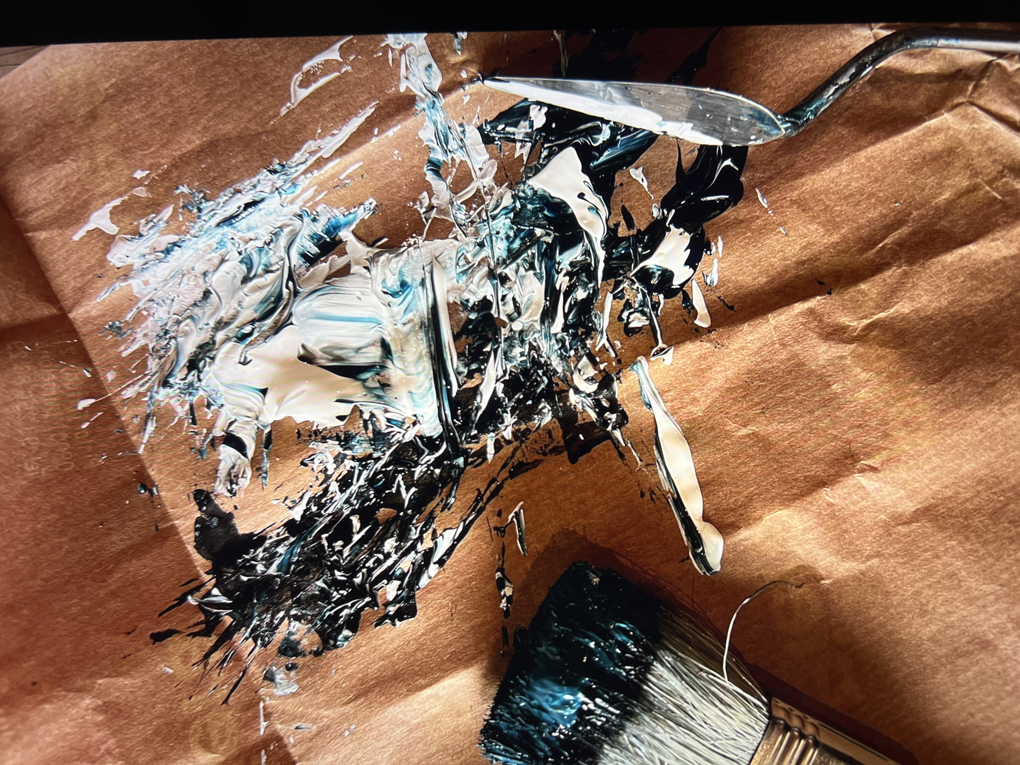 This is a close-up, high-angle shot of art supplies on a textured, brown surface. The surface appears to be paper, possibly kraft paper, with visible wrinkles and folds. A large dollop of thick, white and teal-colored paint is splattered across the center of the image. Swirls of black paint are mixed within the white and teal, creating a dynamic, abstract pattern. To the left, the bristles of a paint brush, heavily loaded with black and teal paint, are visible. To the right, a metal palette knife with a curved handle is positioned, its blade glinting and appearing to have some paint residue on it. The lighting creates strong shadows and highlights, emphasizing the texture of the paint and the paper. The scene depicts an artistic process or the aftermath of one.

The provided image does not contain any recognizable landmarks or details that would allow for its identification within the city of Mollet del Vallès, Spain, or any other specific geographical location.