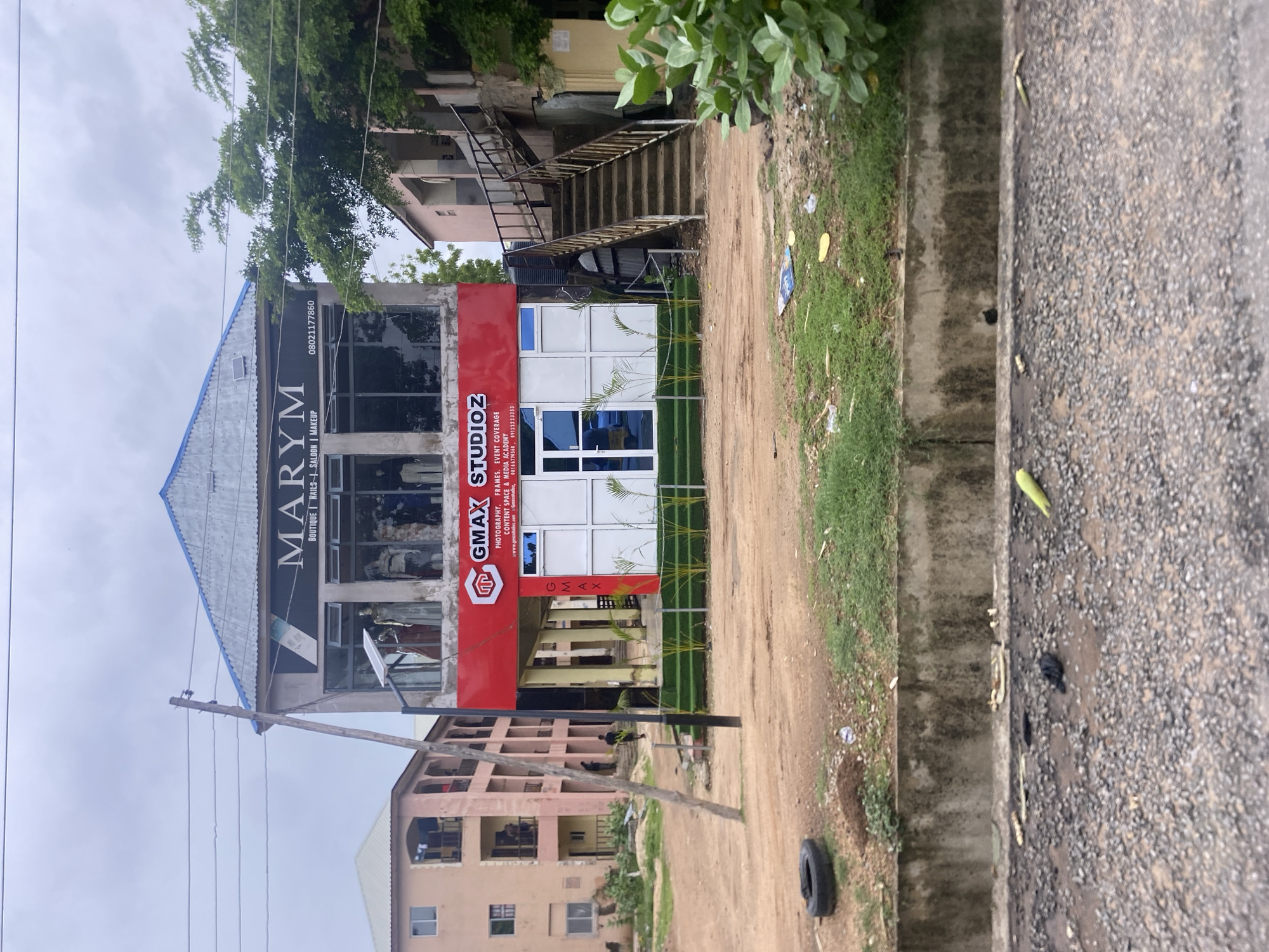 A two-story building stands on a dusty lot in Jalingo, Nigeria. The upper floor of the building houses "MARYM," a business that offers boutique, nails, salon, and makeup services. Several mannequins are visible through the large windows on this floor, displaying clothing. Below, "GMAX STUDIOZ" operates, specializing in photography, frames, event coverage, content space, and a media academy. This lower level features a modern storefront with a bright red facade and glass doors. Palm plants line the front of the GMAX STUDIOZ entrance. To the left of the main building, a multi-story residential building with balconies is visible. To the right, an external staircase leads up to the upper levels of an adjacent building. The scene is framed by a cloudy sky. The ground in front of the building is unpaved, with scattered debris, a discarded tire, and patches of dry grass.
