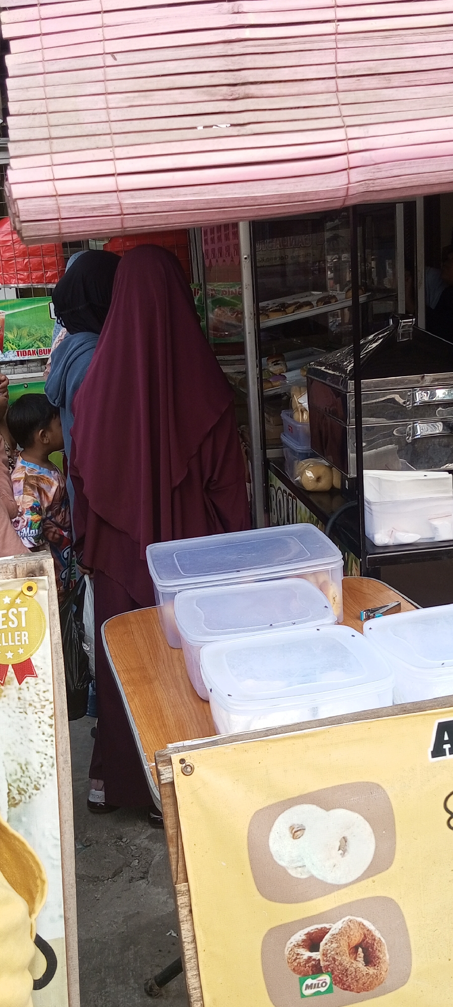In Setu, Indonesia, a street vendor's stall is captured. Three individuals are visible, two women wearing hijabs and a young boy. The woman in the foreground wears a maroon hijab and dress. Behind her, another woman is partially visible in a black hijab and blue jacket. The young boy, wearing a colorful patterned shirt, stands to the left. They appear to be browsing or interacting with the vendor. The stall displays food items in clear plastic containers, with a prominent stainless steel food warmer. A yellow banner with images of donuts is also visible, with the "Milo" logo present on the lower donut image. The background shows a store facade with striped blinds and product displays. The ground is paved with asphalt and debris.