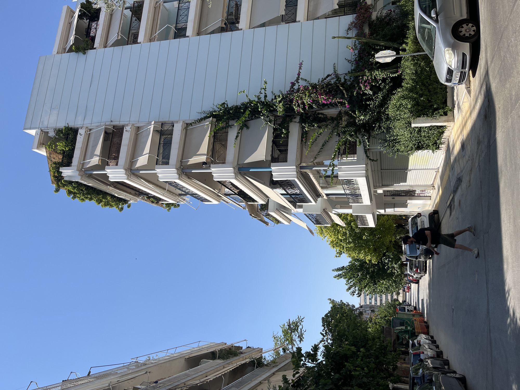 A man is walking across a street in Athens, Greece, on a sunny day. He is wearing a black t-shirt and khaki shorts, with white sneakers. The street is lined with parked cars and buildings. To the right is a tall, light blue apartment building with multiple balconies and awnings. Lush green vegetation and purple bougainvillea vines climb the side of the building. To the left, another apartment building is visible, also with balconies and awnings. Trees line the sidewalk, casting shadows on the street. In the background, a bus and more parked cars are visible, as are other buildings in the distance. The sky is clear and blue.