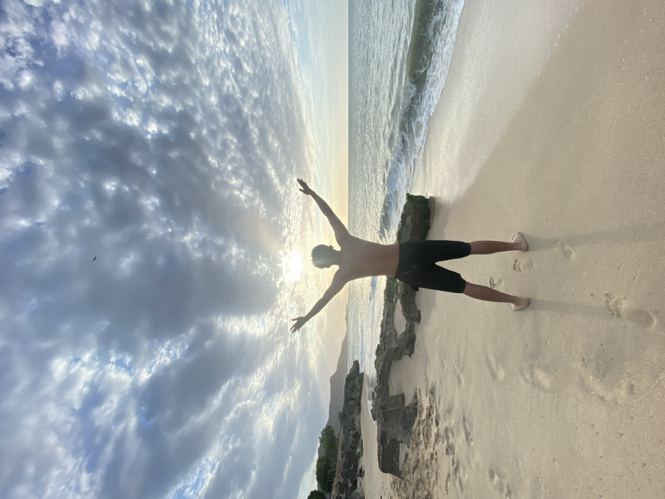 The image displays a sandy beach scene in Juan Griego, Venezuela, at either sunrise or sunset. A lone male individual stands facing away from the viewer, silhouetted against the bright sun positioned low on the horizon. He is bare-chested, wears dark shorts, and has his arms raised and spread wide. The sky above features numerous scattered cumulus clouds, with intense sunlight creating a central glare. To the right, the ocean exhibits gentle waves breaking on the shore. On the left side of the beach, several dark, irregular rock formations are present in the sand. Distant hills with sparse vegetation line the coastline on the far left. Footprints are visible in the light-colored sand near the individual's bare feet. A small, dark avian subject is in flight in the upper left quadrant of the sky.