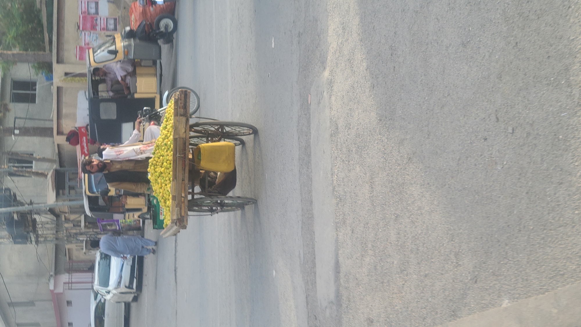 The image depicts a street scene in Quetta City Tehsil, Pakistan. A man stands next to a three-wheeled cart laden with lemons, appearing to be a vendor. Behind him, another man is visible inside a three-wheeled auto rickshaw. Other pedestrians and vehicles are present on the street. The environment appears to be a typical urban setting in Pakistan, characterized by buildings, utility poles, and paved roads.