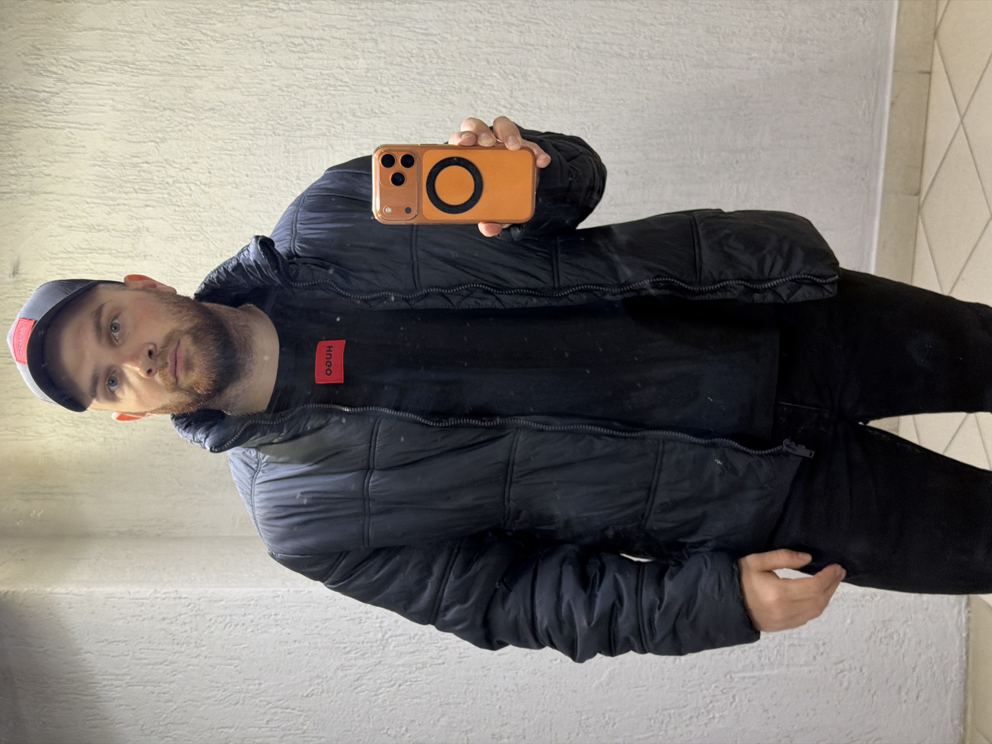 A man with fair skin and a beard is holding an orange cell phone with a black circle on the back. He is wearing a black jacket, a black shirt with a red logo on the collar, and a baseball cap with a red accent. The background consists of a white wall with a textured pattern. The setting is indoors, near what appears to be a corner or edge of a wall with tiled surfaces. The location is Chaikovsky, Russia.