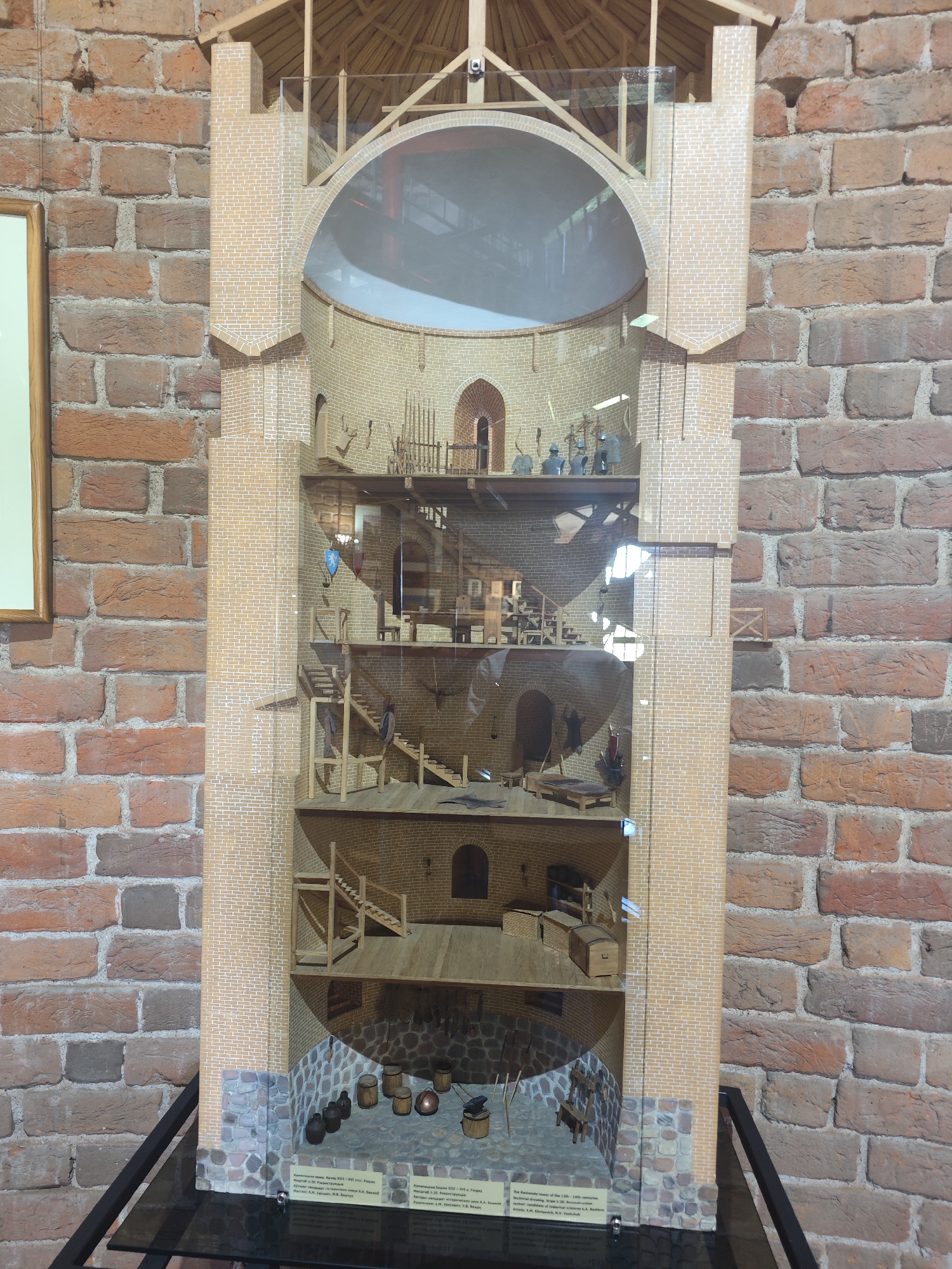 This is a detailed model of a historical brick tower, likely a fortress or defensive structure, situated against a backdrop of a red brick wall. The model is cut in half vertically, revealing multiple interior levels. Each level is furnished with miniature wooden furniture, tools, and human figurines, depicting various activities.
On the lowest level, stone flooring is visible, with several dark, round objects resembling barrels or containers, and a small, dark object that could be a forge or a cooking station. Wooden stairs ascend to the next level, where more barrels and what appear to be wooden tables and chairs are present. Further up, there are more levels with wooden scaffolding, stairs, and various objects. Some levels contain miniature figures dressed in historical attire, engaged in tasks or simply standing. The upper sections of the model show a wooden roof structure.
The exterior of the model is a tall, light-colored brick tower, matching the style of the interior brickwork. The model is encased in glass, and the backdrop is a rough, red brick wall, suggesting it is displayed in a museum or historical exhibition. Below the model, a label with Cyrillic text is visible, indicating the location as "Kamyanyets, Belarus," and the subject as "The Kamyanyets Tower of the 13th-14th centuries." The model appears to be a reconstruction, with a scale indicated as 1:20.
