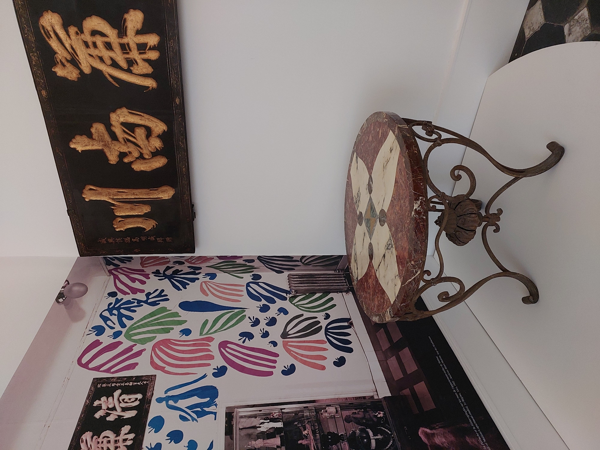 The image shows an interior scene with a round marble-topped table in the foreground. The table has an ornate metal base with scrolled legs and decorative elements. Behind the table, on the wall, is a framed piece of art or signage with gold-colored Chinese characters on a dark background. To the left, there is a large print or mural on the wall featuring colorful, abstract shapes in shades of pink, blue, and green, reminiscent of Matisse's cut-outs. A display cabinet with various figurines is also visible to the left, and a radiator is positioned against the wall. The floor in the background appears to be tiled. The scene is indoors and appears to be a gallery or exhibition space. The location in Nice, France, is identified as the Matisse Museum.
