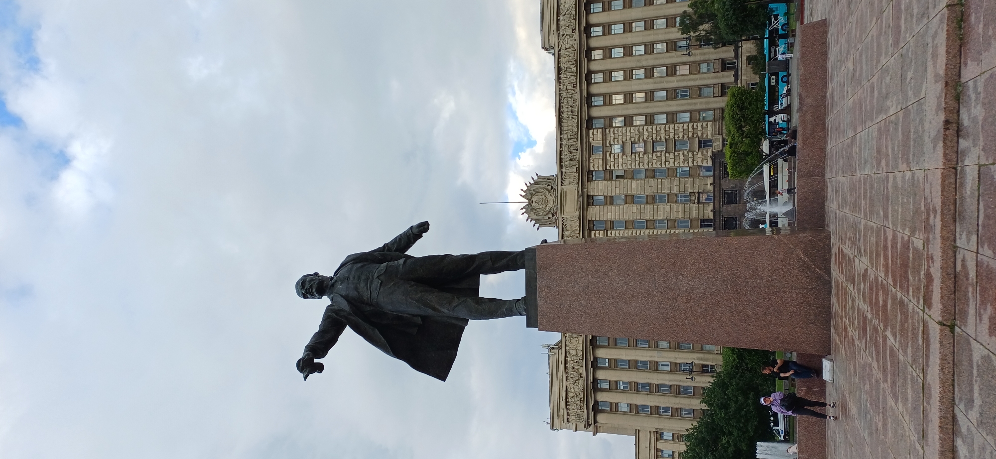 In Saint Petersburg, Russia, a monumental statue of Vladimir Lenin stands prominently in a city square. The statue, cast in dark bronze, depicts Lenin in a characteristic pose, with his left arm extended and pointing forward, and his right arm bent at the elbow, with his hand resting on his chest. He wears a suit and a long overcoat. The statue is mounted on a tall, rectangular pedestal made of polished red granite.
Behind the statue, a grand, multi-story building dominates the background. This building exhibits classical architectural elements, with numerous windows, decorative friezes, and a prominent central crest featuring a hammer and sickle. The building appears to be a significant government or administrative structure, possibly the Smolny Institute, a historic building in Saint Petersburg that has housed various governmental functions.
In the foreground, the square is paved with large, square stone tiles, some of which appear damp, suggesting recent rain or the presence of fountains. Several people are visible in the square. To the left of the statue's base, two individuals are seated on a low ledge. Further left, two women stand, one wearing a purple hoodie and black pants, and the other in dark clothing. In the background, near the building, fountains are active, with water spraying into the air. Buses, some painted in bright blue, are parked or moving in the distance, indicating a public thoroughfare. Trees line the perimeter of the square, adding a touch of greenery to the urban landscape. The sky above is overcast with dramatic clouds, suggesting a diffused daylight.