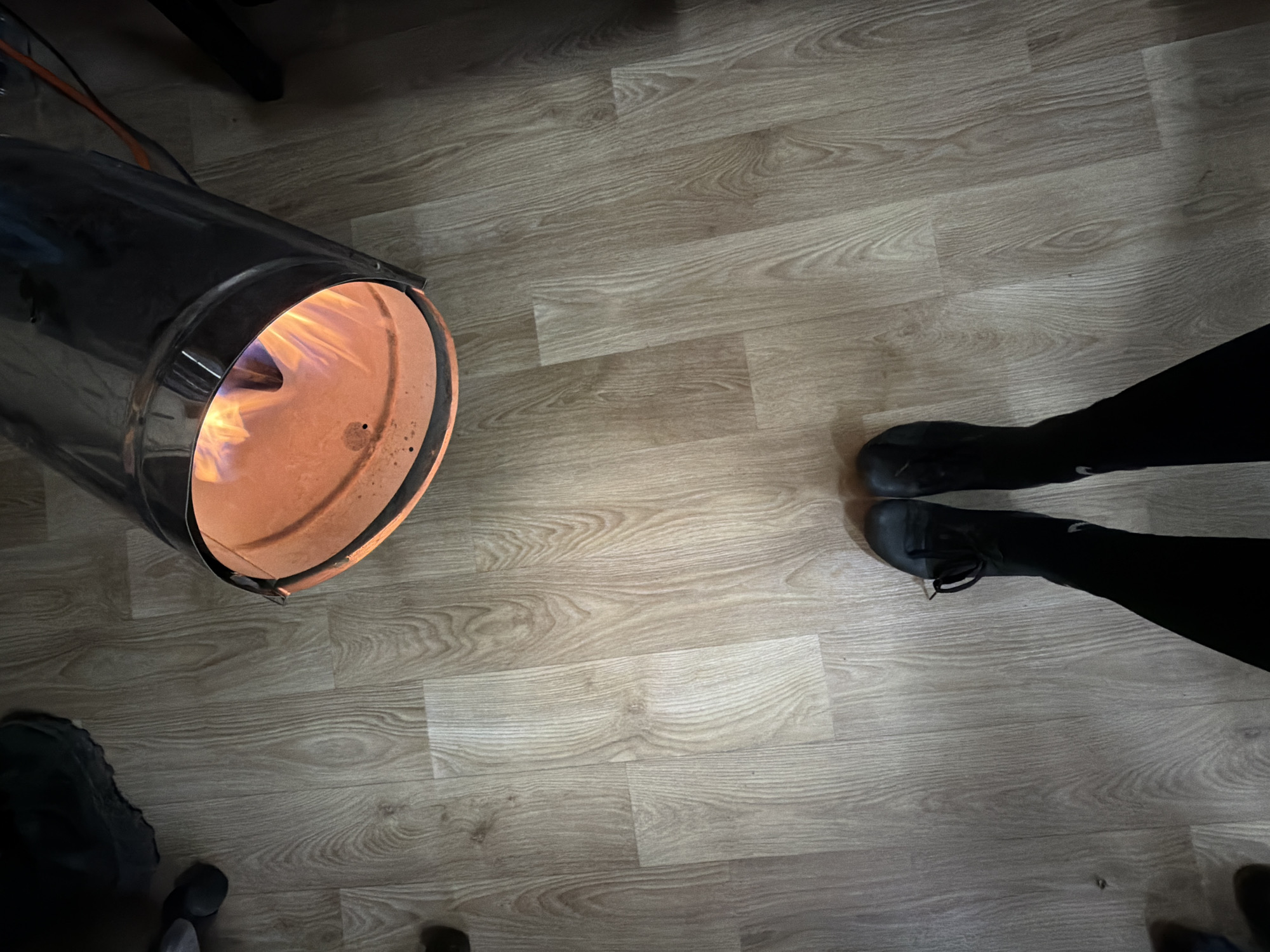An overhead shot shows a lit metal barrel or furnace, two pairs of legs and feet, and a wood-patterned floor. No information is available to place this scene in Plovdiv, Bulgaria. The barrel emits light, possibly flames, suggesting combustion. The person wears black leggings and dark boots. No specific actions or interactions are evident. The environment seems to be indoors with wood flooring.