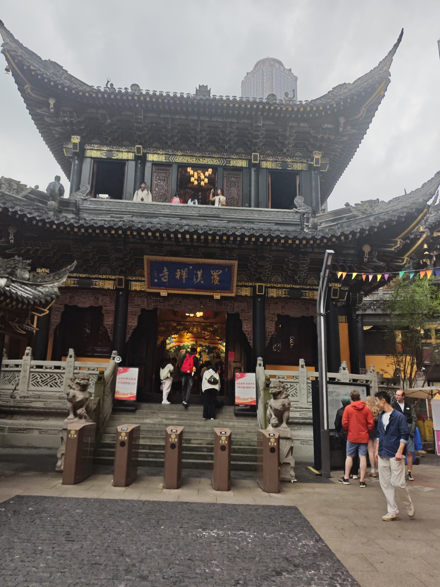 The image depicts a traditional Chinese temple with ornate architecture, located in the Yuzhong District of Chongqing, China. The temple has a multi-tiered roof with intricate carvings and a prominent sign with Chinese characters. People are seen entering and exiting the temple, and some are standing on a balcony observing the scene. Several trash cans and stone lion statues flank the entrance steps. Colorful flags are strung along a side section of the temple. A group of people stand to the side of the temple entrance, one walking away from the building. The foreground shows a paved area, and a modern building is visible in the distance behind the temple. The sky is overcast, suggesting a cloudy day.