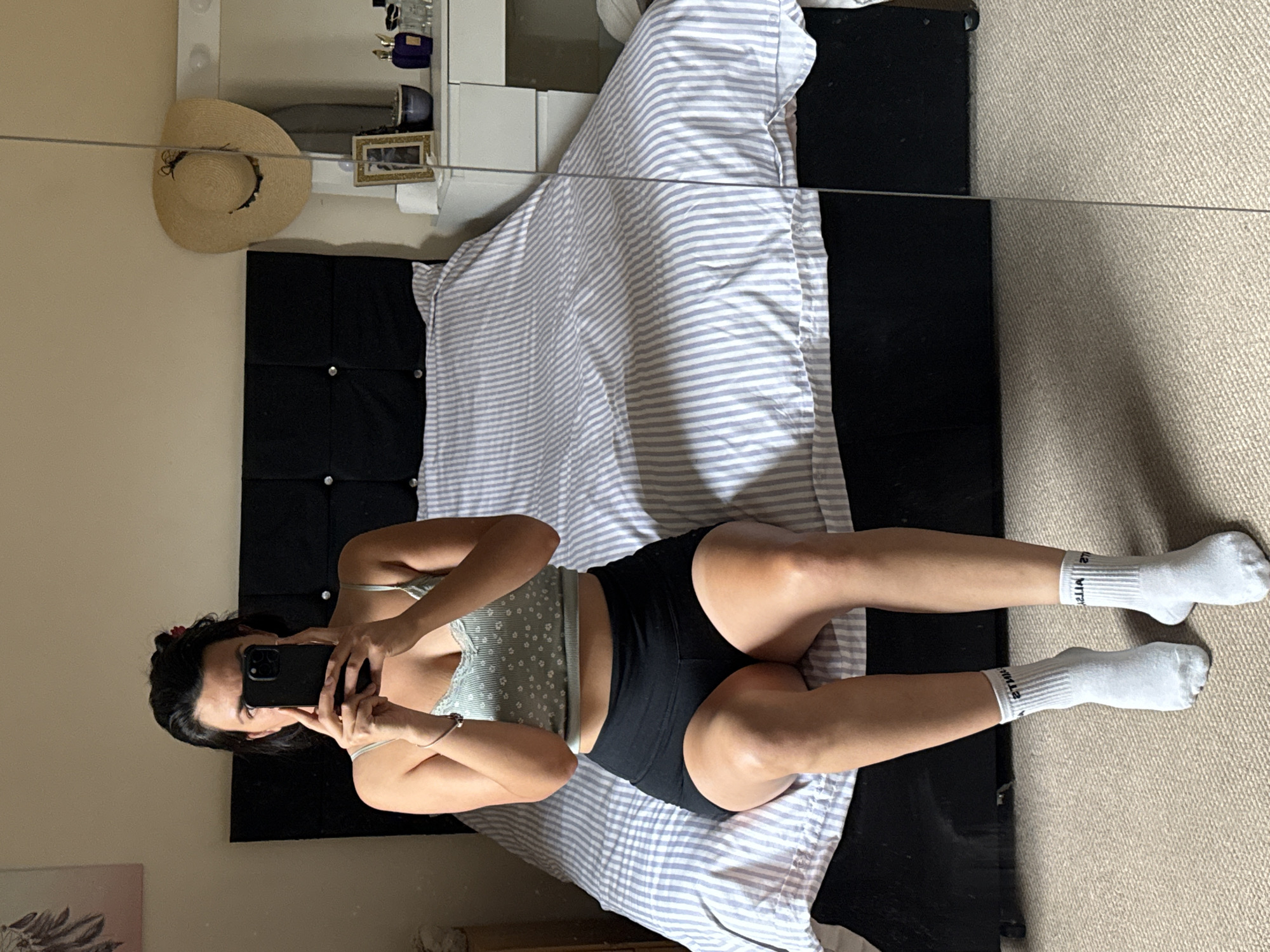 A full-body shot shows a woman sitting on the edge of a bed. She is holding a smartphone up to take a picture of herself. She is wearing a light green crop top with a floral pattern and black athletic shorts. She also has on white crew socks. The bed has a black headboard and is covered with a duvet that has blue and white stripes. A large mirror is positioned to the right of the bed, reflecting part of the room. The room appears to be a bedroom, with a dresser visible in the background. The floor is carpeted in a light beige color. The image was taken in Great Yarmouth, United Kingdom.