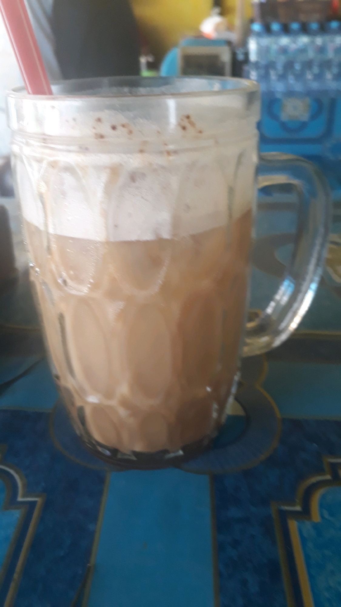 An extreme close-up, vertical shot focuses on a clear glass mug filled with a light brown, frothy beverage. The mug has a distinct textured pattern of raised circles around its body and a large handle on the right side. Two pink straws extend from the top of the drink. The beverage itself appears to have layers, with a lighter, foam-like substance at the top, dotted with small brown specks, and a denser liquid below. The mug sits on a surface that appears to be a table covered with a blue tablecloth featuring an intricate pattern of gold and white designs. The background is blurred and out of focus, showing hints of a yellow wall, some blue plastic bottles, and possibly a dispenser. The lighting is somewhat dim, with a slight glare on the glass. The image composition is tight, with the mug occupying most of the frame.