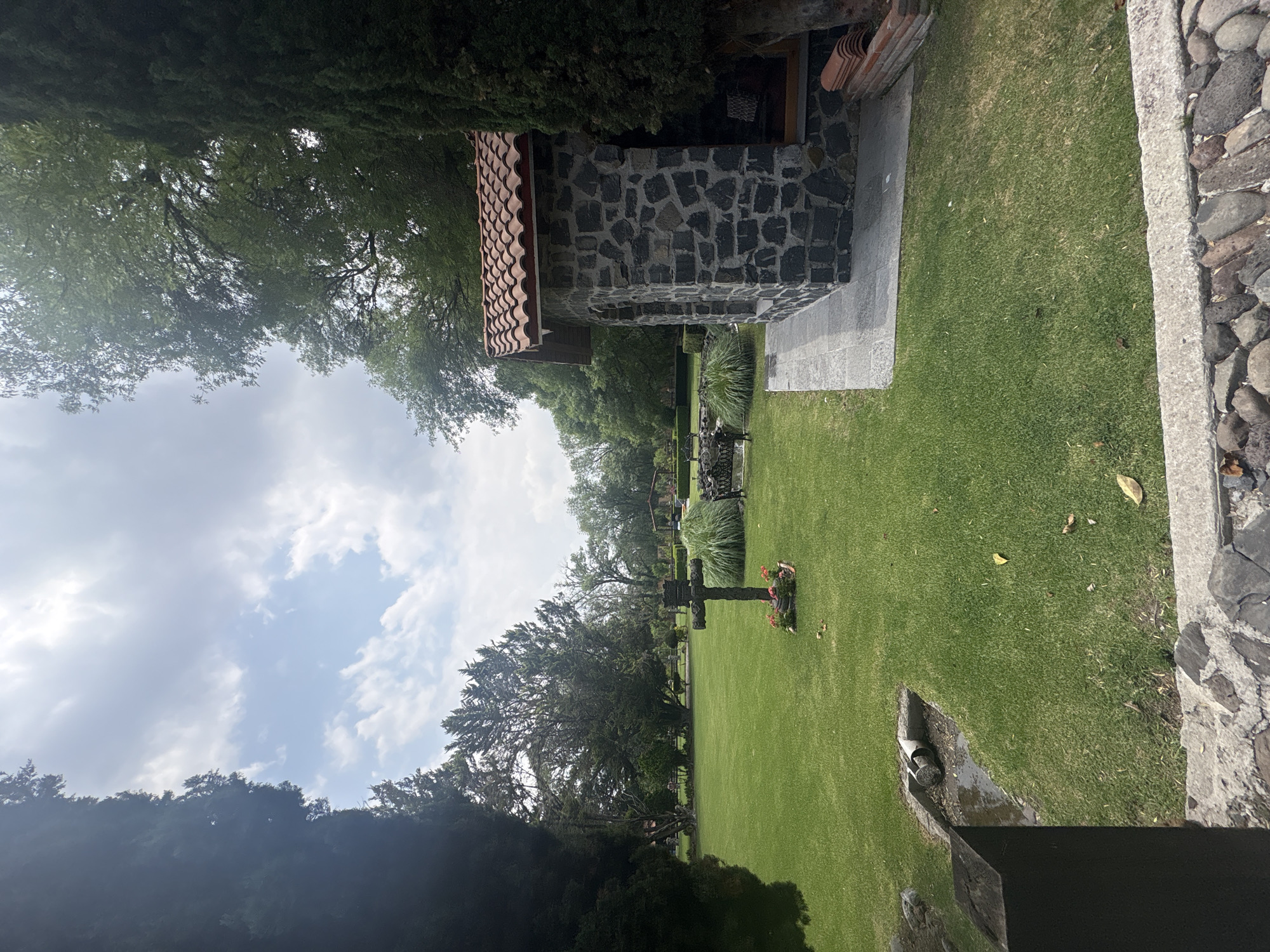 The image depicts a cemetery landscape in Cañada de Madero, Mexico. A stone building with a tile roof stands to the side of a green lawn dotted with headstones and crosses. One notable cross is visible in the center, adorned with flowers. Trees surround the perimeter, with a partly cloudy sky above.