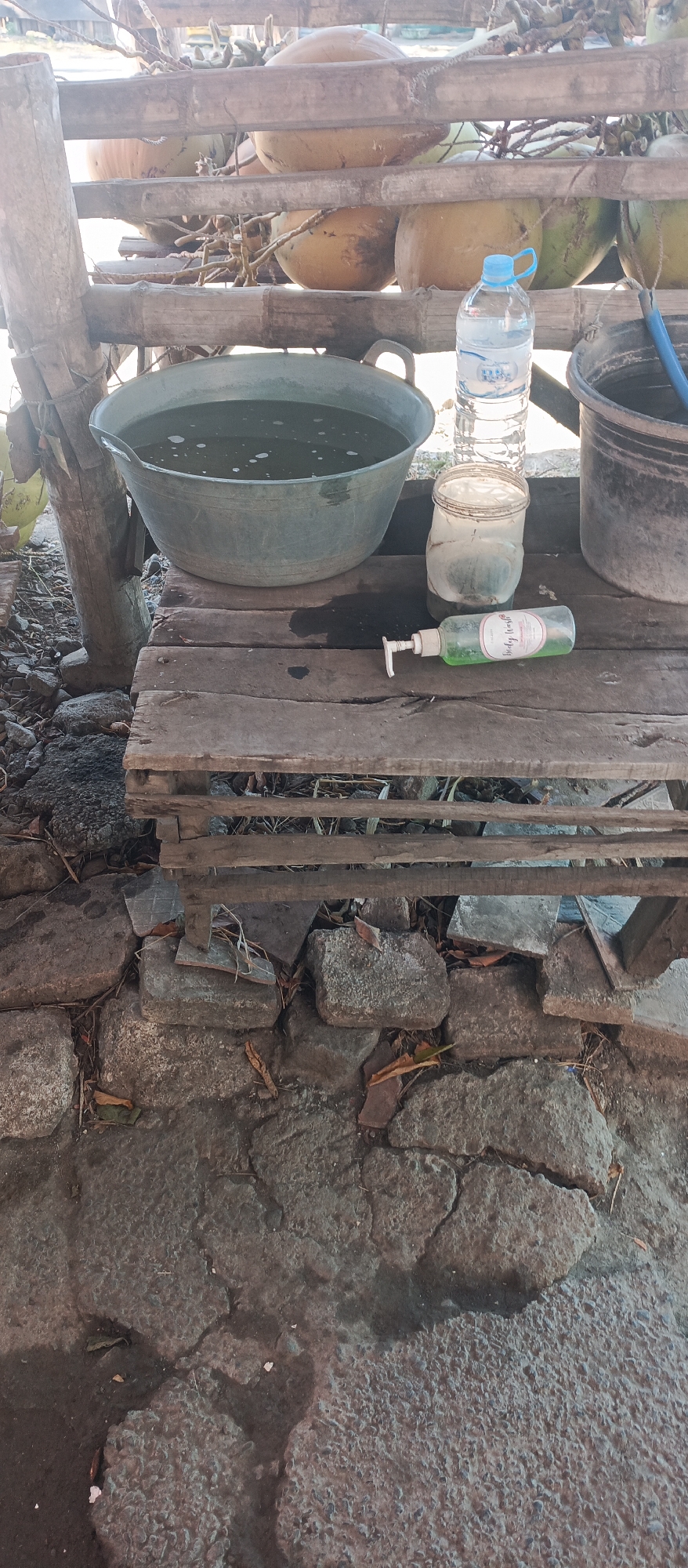 The image depicts an outdoor scene in Jatisari, Indonesia, with a focus on a rustic wooden surface supporting several objects. A large, gray basin filled with dark liquid and bubbles sits to the left. Beside it, a clear plastic water bottle with a blue cap is upright, and next to that is a clear plastic jar with a lid that appears to contain a green substance. A green pump bottle labeled "Body Wash" lies horizontally in front of the jar. To the right of the jar is a dark bucket. The wooden surface itself is composed of planks of varying widths and shows signs of age and wear. In the background, a bamboo structure supports large, round objects, possibly coconuts. The ground beneath the wooden table is a rough, textured surface composed of stones and scattered dry leaves.
