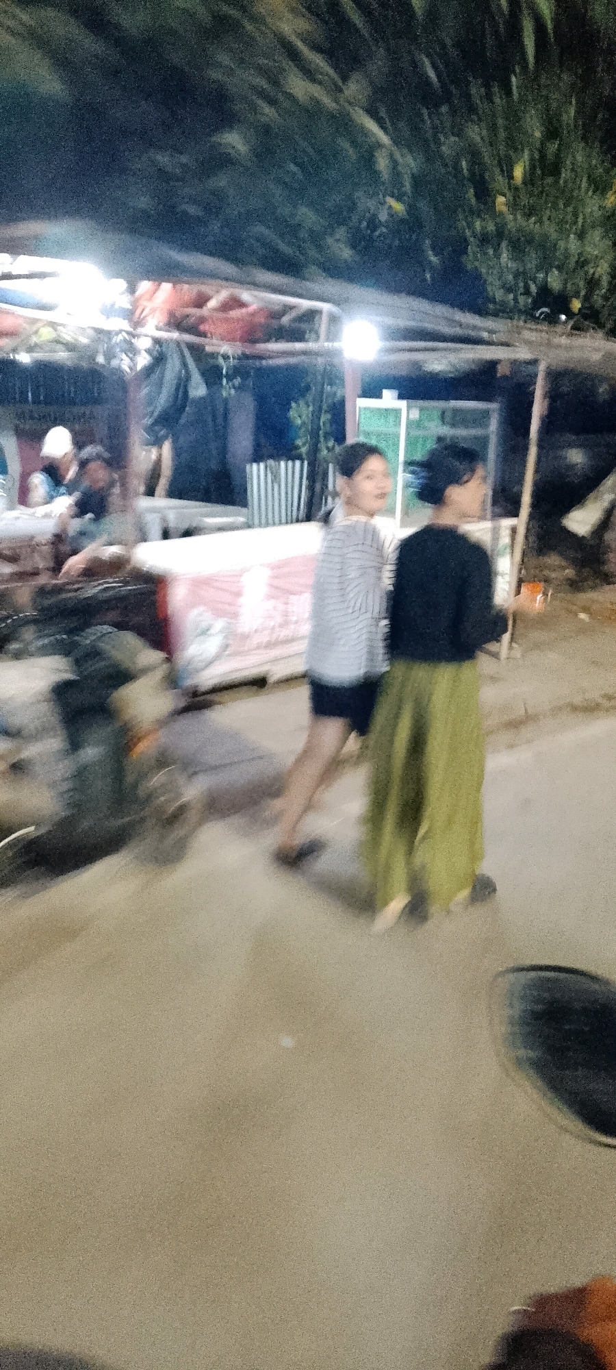 The image captures a nighttime scene on a street, likely a market or roadside stalls, in Bekasi, Indonesia. Two women are the primary subjects, walking from left to right across the frame. The woman on the left wears a horizontally striped long-sleeved shirt and dark shorts. She has dark hair pulled back and is looking towards the left. The woman on the right is dressed in a dark long-sleeved top and a long, flowing olive-green skirt. Her hair is also dark and pulled back. She appears to be looking to her right.
The environment is dimly lit, with artificial lights illuminating the scene. To the left and in the background, there are structures that appear to be temporary stalls or shelters, possibly with goods displayed. One stall has red items visible. In the middle ground, there are what seem to be tables or platforms, one covered with a white and pink patterned cloth. The ground is paved with a light-colored material, possibly asphalt or concrete, showing some texture and variations in tone. There is a noticeable motion blur throughout the image, suggesting movement of the subjects or the camera. In the lower left corner, a dark, circular object, possibly a wheel or a tire, is partially visible. The overall impression is a bustling, informal commercial area at night.