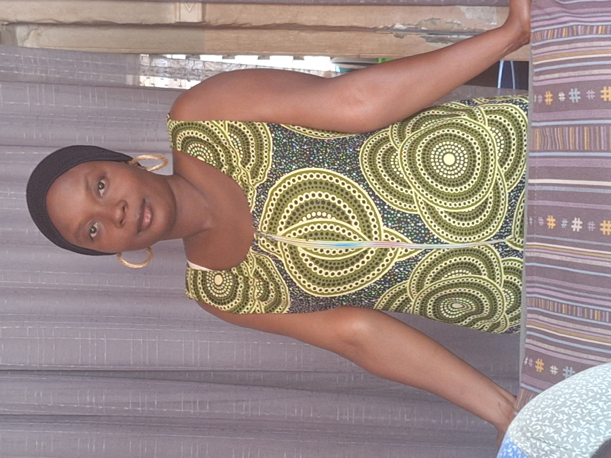 A woman is indoors, posing for a photograph. She is wearing a dress with a green, black, and white circular pattern and large gold hoop earrings. She also has a black head covering. The background includes a gray curtain and a patterned striped fabric. It is unclear if the location is indeed Sabon Tasha, Nigeria, as this cannot be verified from the visual information.