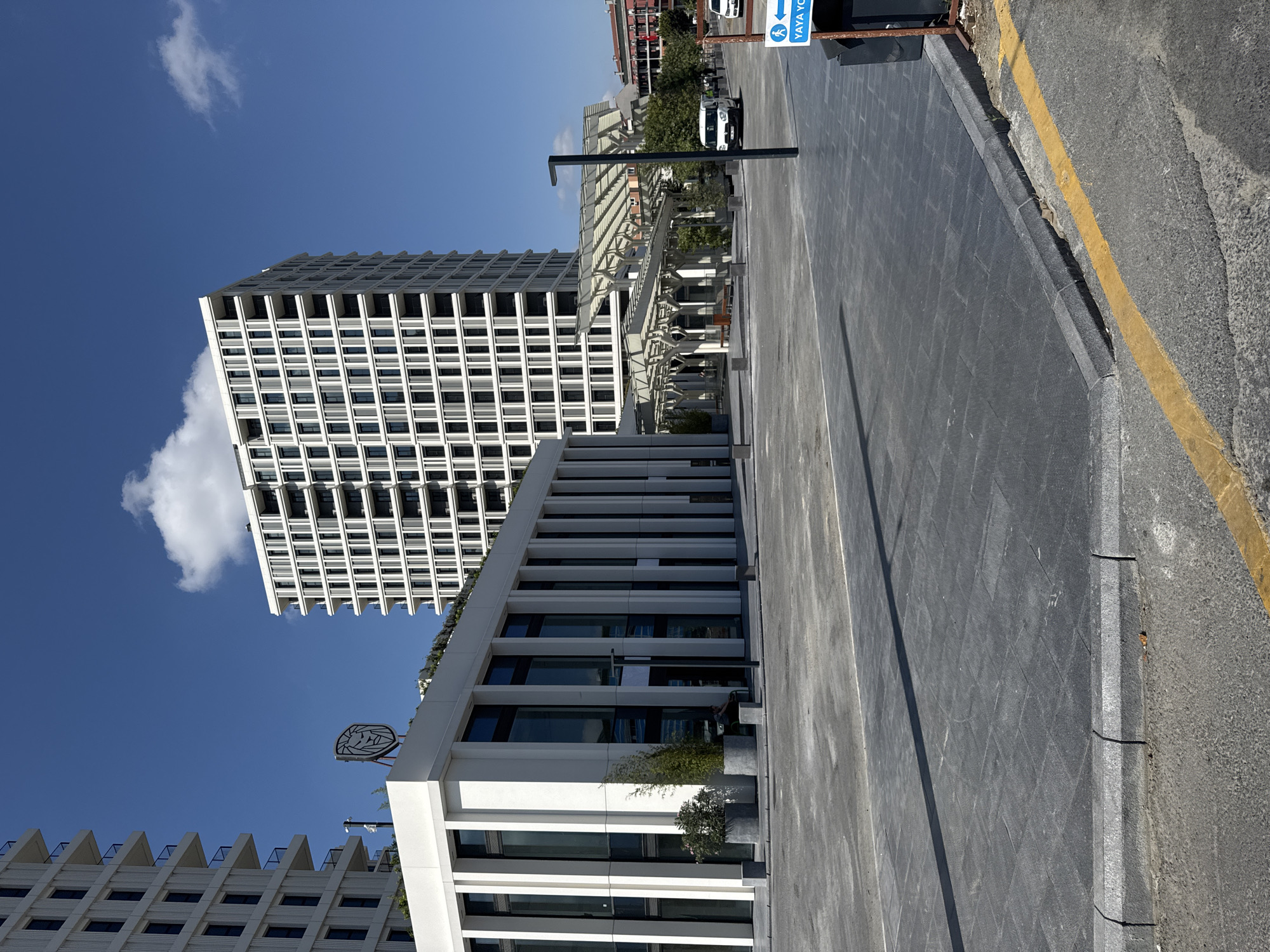 The image shows a modern urban scene with a clear blue sky and scattered clouds. In the foreground, a paved area with dark grey square tiles transitions to a darker asphalt surface with a yellow painted line along the curb. To the right, a dark blue sign with white text indicates a pedestrian path.
Dominating the midground are several contemporary buildings. To the left, a white, multi-story building with vertical lines and dark-tinted windows is partially visible. On its roof, a prominent sign featuring a stylized lion's head logo is mounted. Adjacent to this building is a lower, white structure with a colonnaded facade of vertical white elements and large, dark windows. Planters with greenery are situated outside this building.
In the background, a tall, white high-rise building with a grid-like pattern of windows and balconies stands prominently. Another building with red accents is visible further in the distance. Between the midground and background buildings, a covered walkway or canopy structure with a white, skeletal design extends across the scene. A white van is parked in front of this structure. A person is seated on a bench in the shadow of the covered walkway. The environment appears to be a newly developed urban area, possibly a commercial or residential complex. The location is identified as Bahçelievler, Turkey.