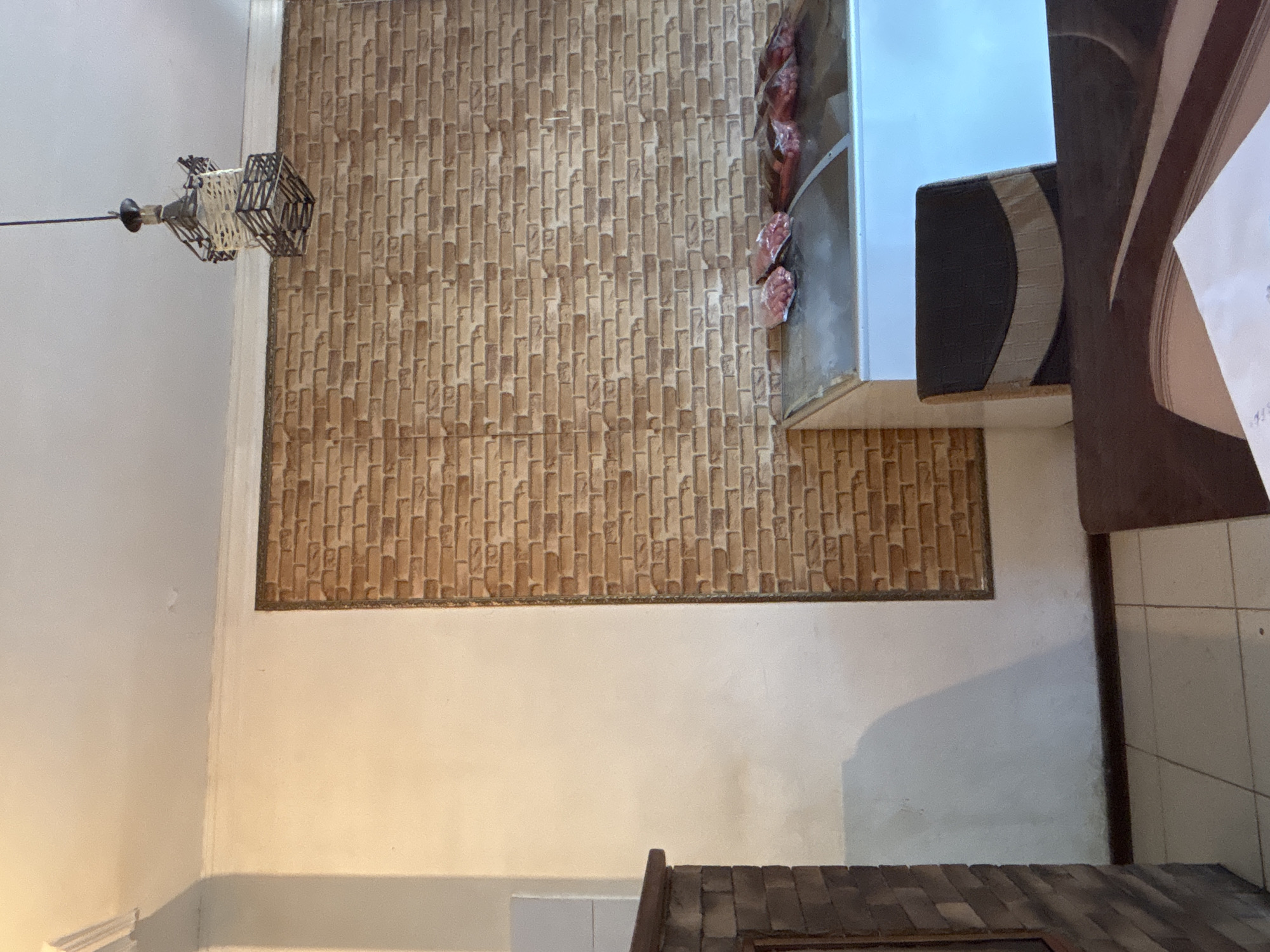 The image shows a dimly lit room with a brick-patterned wall as a prominent feature. A metallic pendant light hangs from the ceiling on the left side of the frame. On the right, a white cabinet or shelf is visible, containing what appear to be small, packaged food items, possibly meat. The cabinet's edge is dark and curved. Further to the right, a dark wooden surface with a patterned inlay is visible, and a piece of paper with some writing is partially visible on it. The lighting suggests it might be evening or indoors with artificial light. The scene appears to be a domestic or commercial interior, possibly a kitchen or pantry area. There is no indication of the time of day or weather. The overall impression is one of a still life arrangement, with no people or active events depicted. The brick pattern on the wall is a repeating motif of small, rectangular blocks in shades of brown and tan. The white walls contrast with the darker elements of the scene.