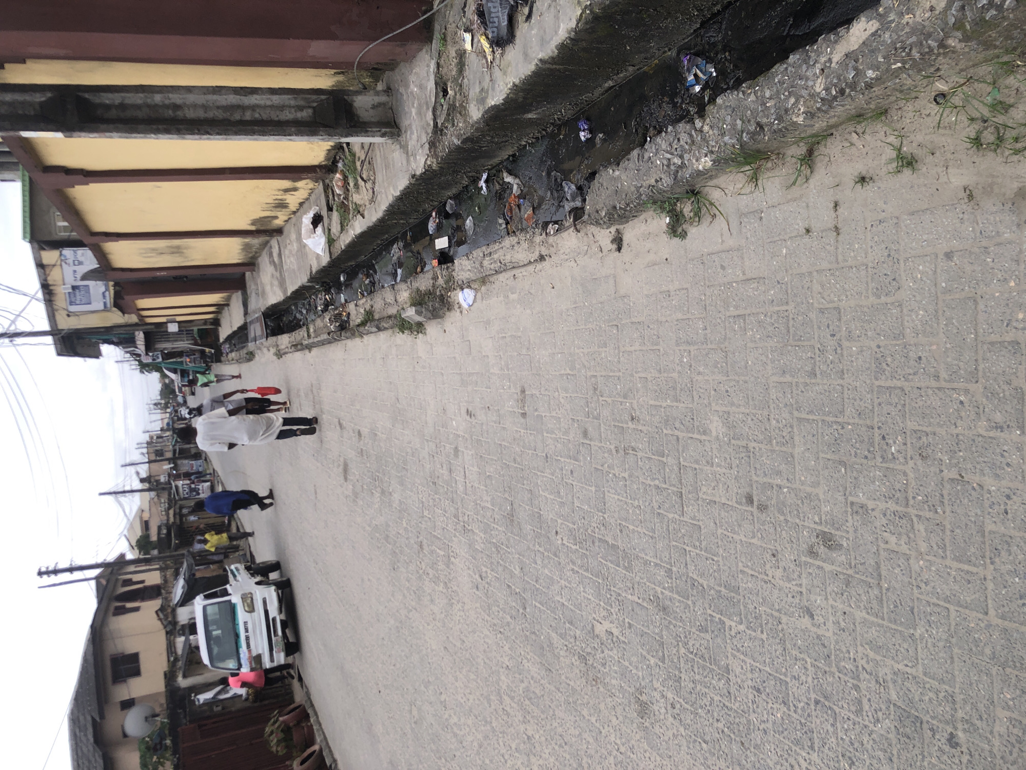This is a street-level photograph of a residential area in Ado, Nigeria. The scene depicts a paved street with a drainage channel running alongside it on the right. The drainage channel is filled with dark, stagnant water and is littered with various types of refuse, including plastic bags, bottles, and other debris.
On the left side of the street, several people are visible walking. One person, dressed in white, is walking away from the camera down the street. To the left of the road, a white pickup truck is parked, with a woman in a pink top standing near it. Further down the street, other individuals are seen walking, some of them carrying red bags. The buildings lining the street appear to be residential or small commercial establishments, with a large yellow wall with a red border dominating the right side of the image, adjacent to the drainage channel. The sky is overcast, suggesting a cloudy day. The pavement of the street is made of interlocking bricks.