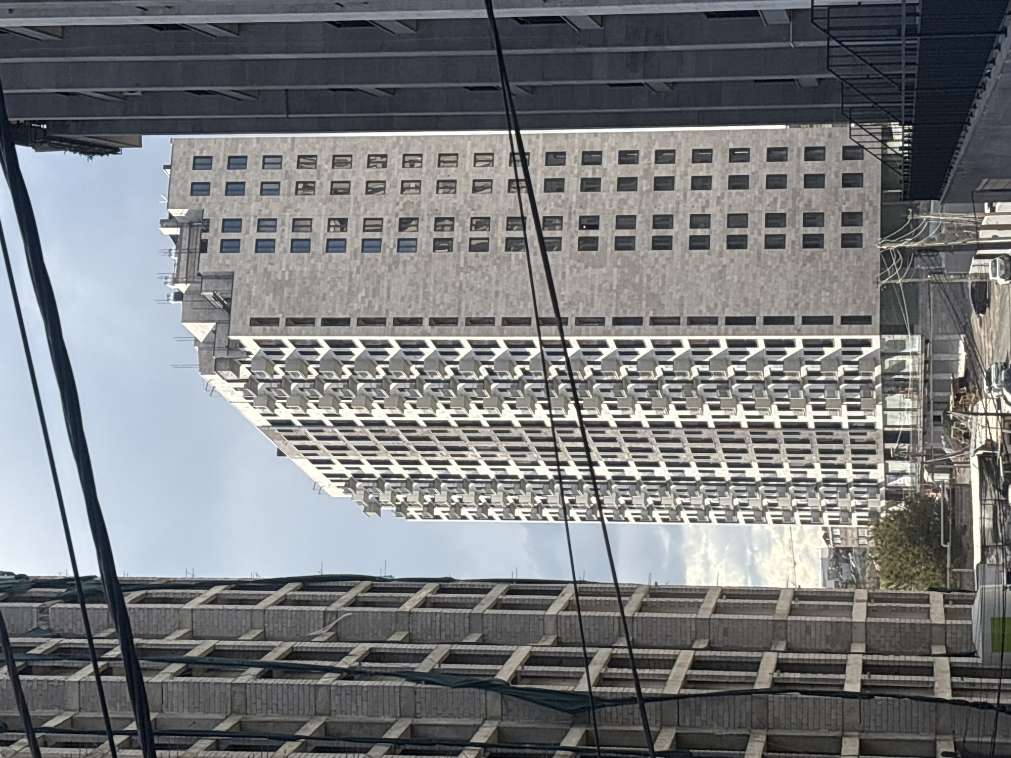 The image shows a tall, light grey building with numerous windows, viewed from a low angle, framed by sections of other buildings and overhead wires. The building has a distinct architectural style with rows of projecting balconies below the windowed section. Below the building in the foreground is another building that appears to be under construction or renovation with exposed concrete framework. The sky is partially visible in the background, between the buildings. The image was taken in Yerevan, Armenia.