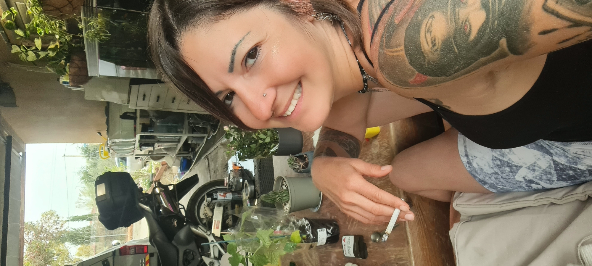 A woman with a nose piercing and tattoos on her arm is taking a selfie while holding a cigarette. She is smiling and looking directly at the camera. In the background, there is a motorcycle, several potted plants, and a parked car. The location appears to be a garage or outdoor workspace, possibly in the Lachish Regional Council area of Israel.