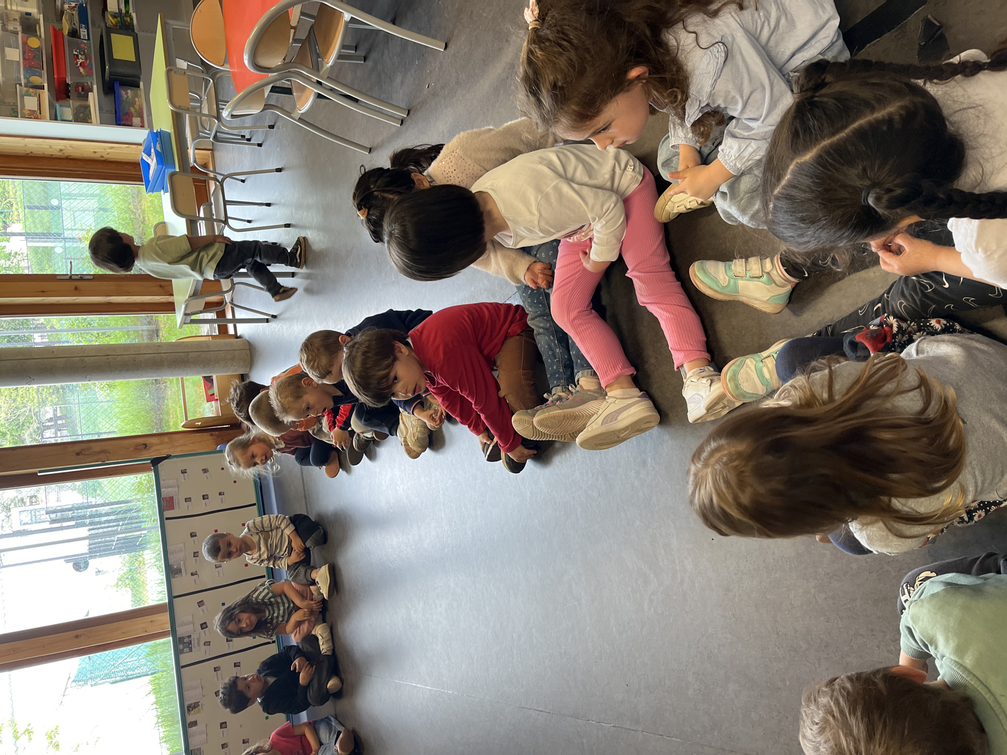 A classroom in La Fouillouse, France, features approximately 20 children of preschool age seated on the floor in a semi-circle. The children are of varying ethnicities, and they are dressed in casual clothing. Their attention is focused downward, possibly on an object or activity in the center of the semi-circle. To the left of the group, a row of low storage units is visible. Natural light enters the room through a window on the left side. Chairs and tables are arranged in the upper-left corner of the frame. One child stands near the window. The floor is a neutral gray color.