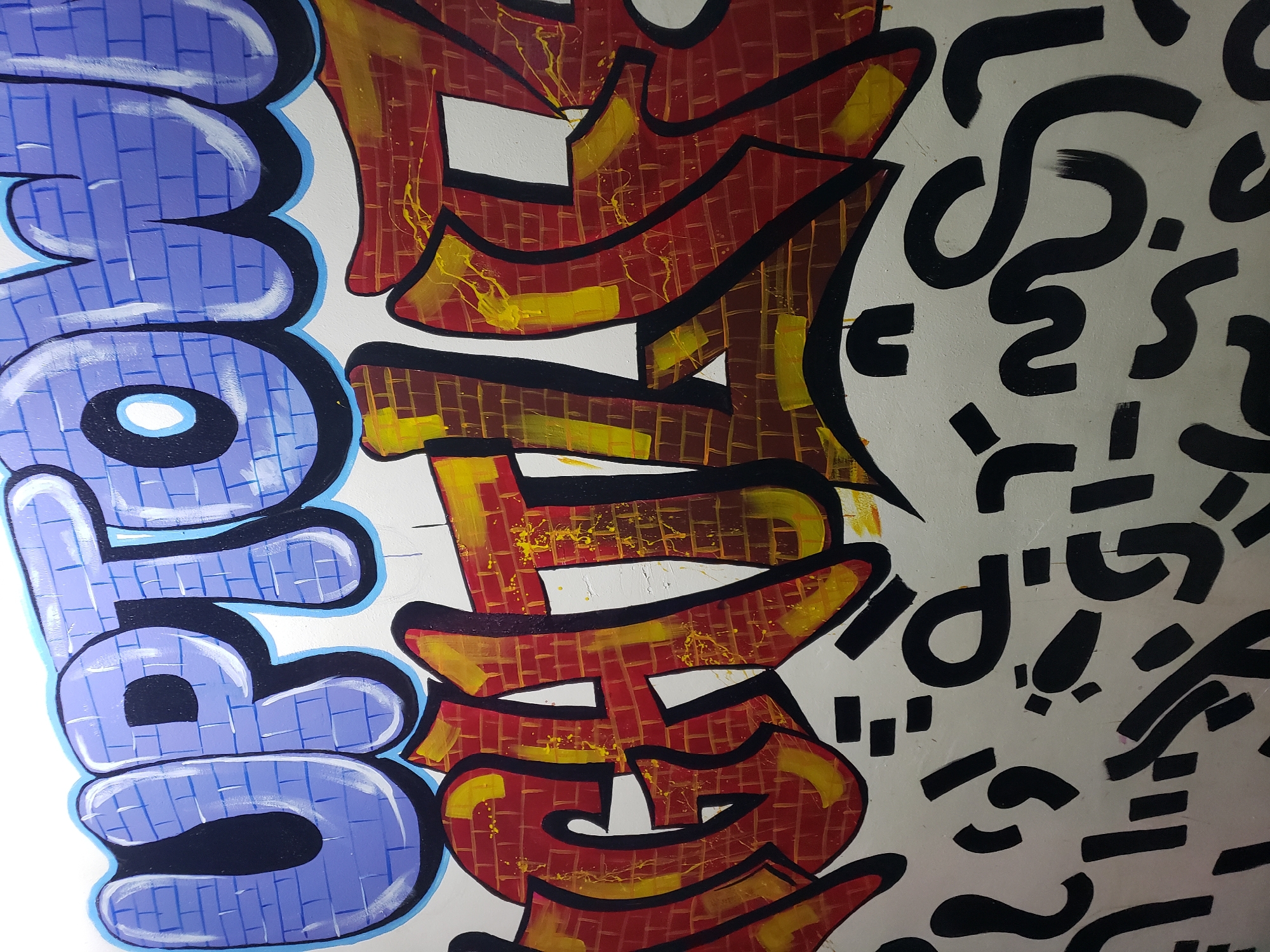The image presents a section of graffiti art on a wall, likely in an urban environment, possibly in Adankolo, Nigeria. On the left side, a word in large, stylized blue bubble letters with a white fill and a black outline is visible. The word is oriented vertically. Adjacent to it is another stylized word in red and yellow, textured like brickwork, with black outlines and white spaces within the letters. To the right of the red and yellow word, the background is white and covered with randomly oriented black shapes resembling letters, symbols, or abstract forms. The style suggests a contemporary art piece or mural. The photograph is taken at an angle, looking upwards.