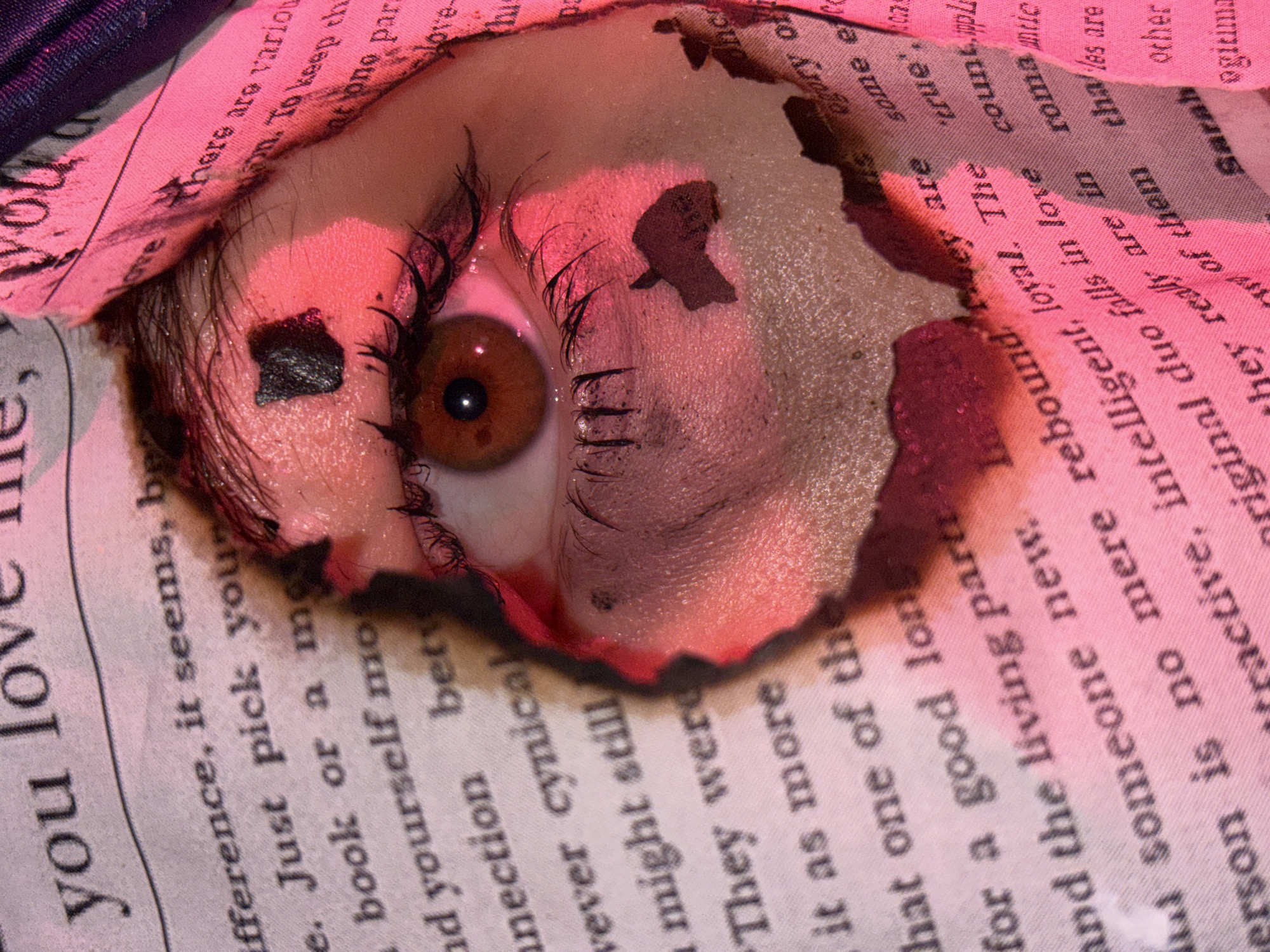 This close-up image presents a human eye intently peering through a jagged, burned opening in a piece of paper or fabric. The eye has a warm brown iris and is framed by visible eyelashes and eyebrow hairs, some of which appear singed or smudged with dark residue, possibly ash or makeup. The skin around the eye is bathed in a dramatic pinkish-red glow, suggesting a direct, colored light source. Scattered dark, charred fragments rest on the skin and around the eye.
The material surrounding the eye is a light-colored paper or fabric, heavily textured and printed with black serif text. The edges of the opening are conspicuously burned and frayed, displaying tones of orange and deep red where the material has been charred. The visible text is fragmented and includes phrases such as "you love," "difference, it seems, be," "Just pick your," "book or a mo," "and yourself mo," "connection bet," "cynical," "might still," "They were," "it as more," "for a good lon," "and the living part," "someone new," "There are variou," "To keep th," "love," "ogory of," "tractive, intelligent, loyal. The true," "original duo falls in love count," "them that are," "Sarah," and "ogiuma."
The overall scene conveys an intense, introspective, or perhaps unsettling mood, with the eye seemingly observing or emerging from a context of damage or revelation. The lighting is artificial, making the time of day indeterminate. This staged shot, set in Perm, Russia, appears to be an artistic commentary on perception or resilience amidst destruction.