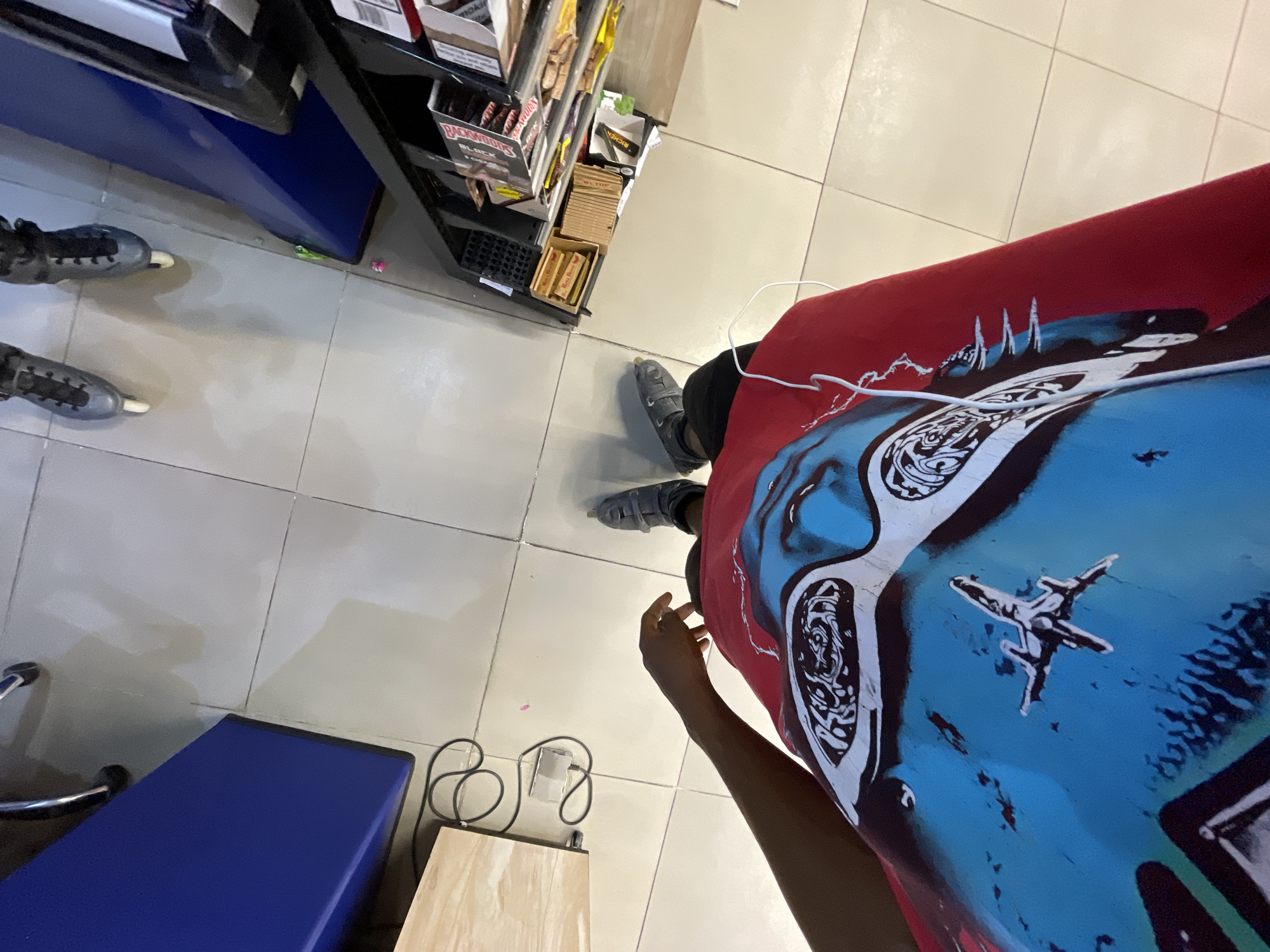 A high-angle, full shot captures a person wearing rollerblades and a red t-shirt with a graphic design of a fighter jet and a stylized face. The person's left arm is visible. The background shows a tiled floor with what appears to be shelves or displays of products, a blue cabinet, and a wooden table with a coiled black cord and a smartphone. The scene is likely an indoor retail environment in the city of Moba, Nigeria.