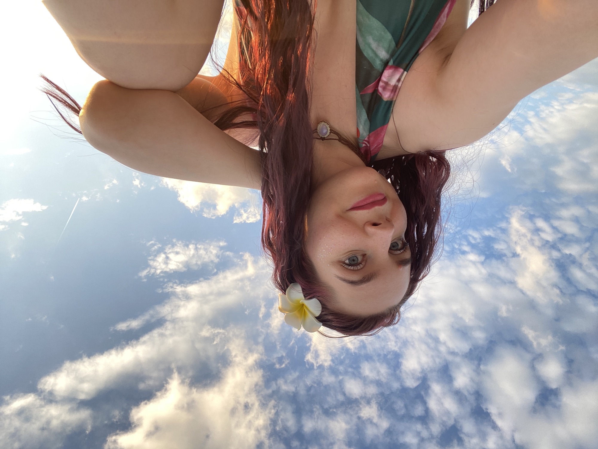 A woman is positioned in the foreground, looking directly at the camera. She has fair skin, red hair, and is wearing a flower in her hair. The background is a blue sky filled with clouds.
It is not possible to determine the location in the city of Chaikovsky, Russia, from this image.